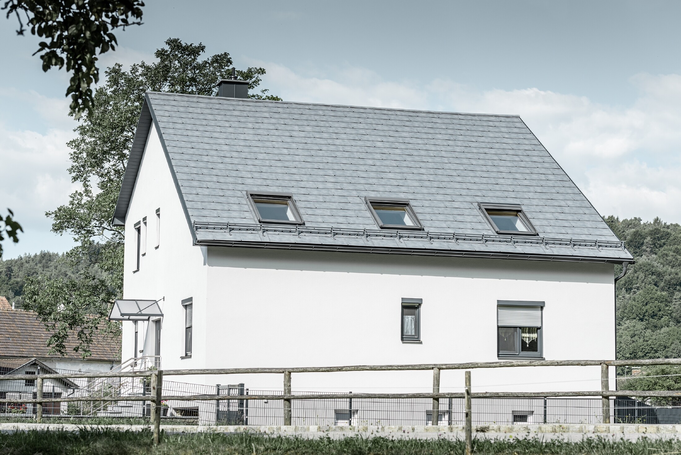 Dach dwuspadowy klasycznego domu jednorodzinnego został pokryty nową dachówką klasyczną R.16 PREFA w kolorze szarym kamiennym. W połaci dachowej wkomponowano trzy okna dachowej i zamontowano bariery śniegowe. Elewacja jest utrzymana w kolorze białym.