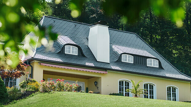 Dom jednorodzinny ze zmodernizowanym dachem z dachówką PREFA w kolorze antracytu z zaokrąglonymi oknami dachowymi (wole oko) i białym kominem.