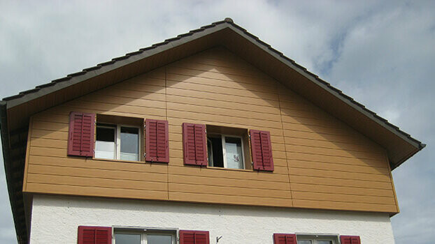 Okładzina ściany szczytowej klasycznego domu z dachem szczytowym. Ściana szczytowa jest obłożona ułożonymi poziomo sidingami PREFA w kolorze dąb naturalny. Okna mają czerwone okiennice.
