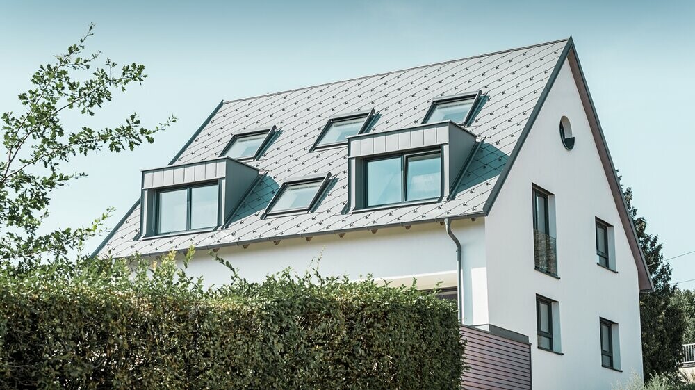 Renowacja dachu dwuspadowego z dwiema lukarnami i czterema oknami dachowymi. Dach został pokryty dachówką rombem PREFA 44 × 44 w kolorze jasnoszarym. Rynna wisząca PREFA także jest w kolorze jasnoszarym. 