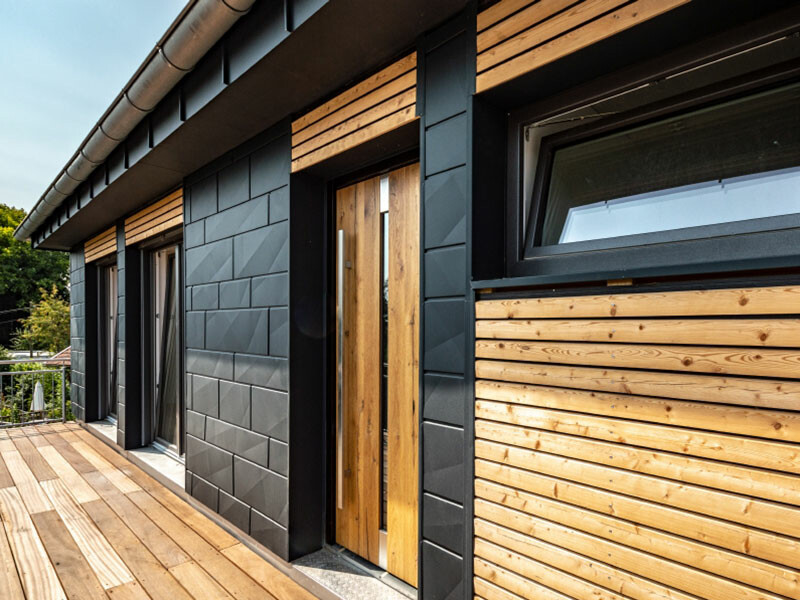 Nowoczesny dom jednorodzinny z pięknymi panelami elewacyjnymi PREFA Siding.X w kolorze antracytowym i elementami z drewna.