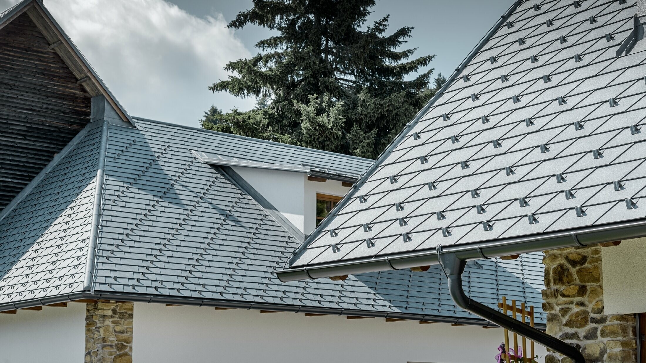 Detal aluminiowego krycia dachu PREFA, dachówka łupkowa w kolorze szarym kamiennym z aluminiową rynną dachową PREFA w kolorze antracytowym; w tle jest widoczna lukarna ciągniona z dachem na rąbek stojący. Elewacja jest biała z osadzonymi elementami kamiennymi.