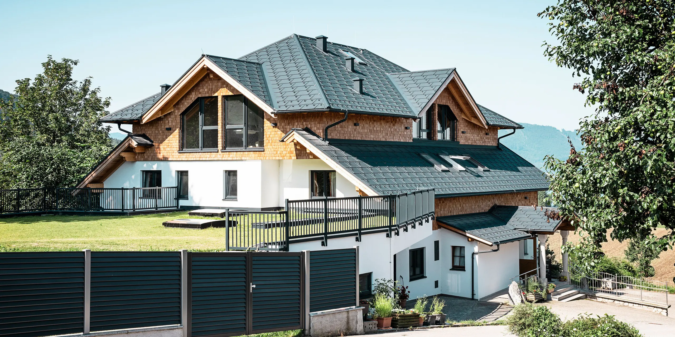 Seitenansicht des Café-Restaurants Zenz'n Stub'n in Schörfling am Attersee mit modernem Aluminiumdach aus PREFA Dachplatten R.16 in der hochwertigen Beschichtung P.10 Anthrazit. Das langlebige PREFA Dach vereint Ästhetik und Funktionalität und bietet optimalen Schutz. Der Zugang zum Garten und die harmonische Einbettung des Gebäudes in die natürliche Umgebung unterstreichen das zeitlose Design.