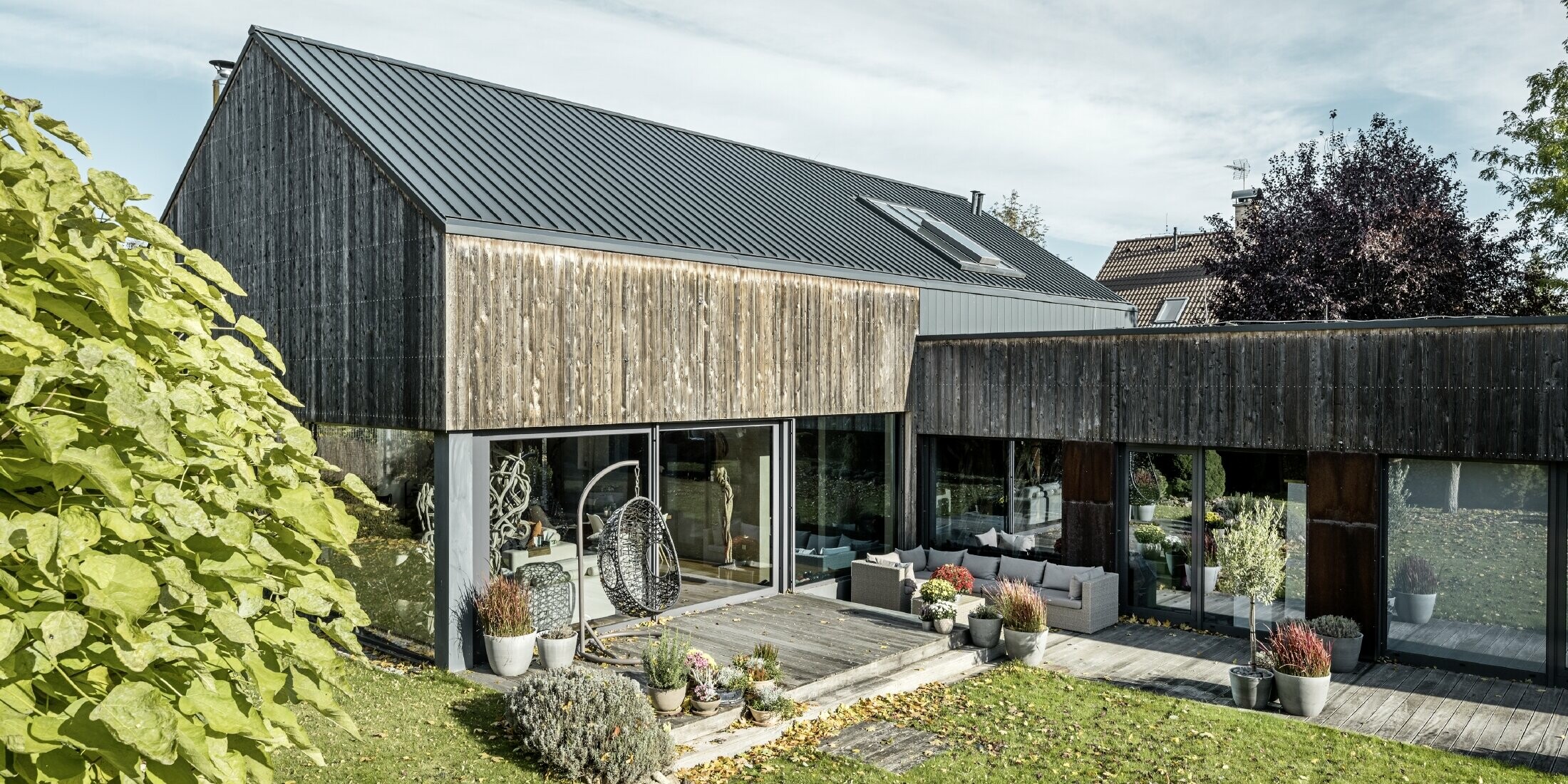 Dom jednorodzinny z dachem dwuspadowym, pokryty PREFALZ w technologii rąbka podwójnego w kolorze antracytowym z elewacją z postarzanego drewna. Z pięknym tarasem drewnianym i dużymi oknami na parterze.