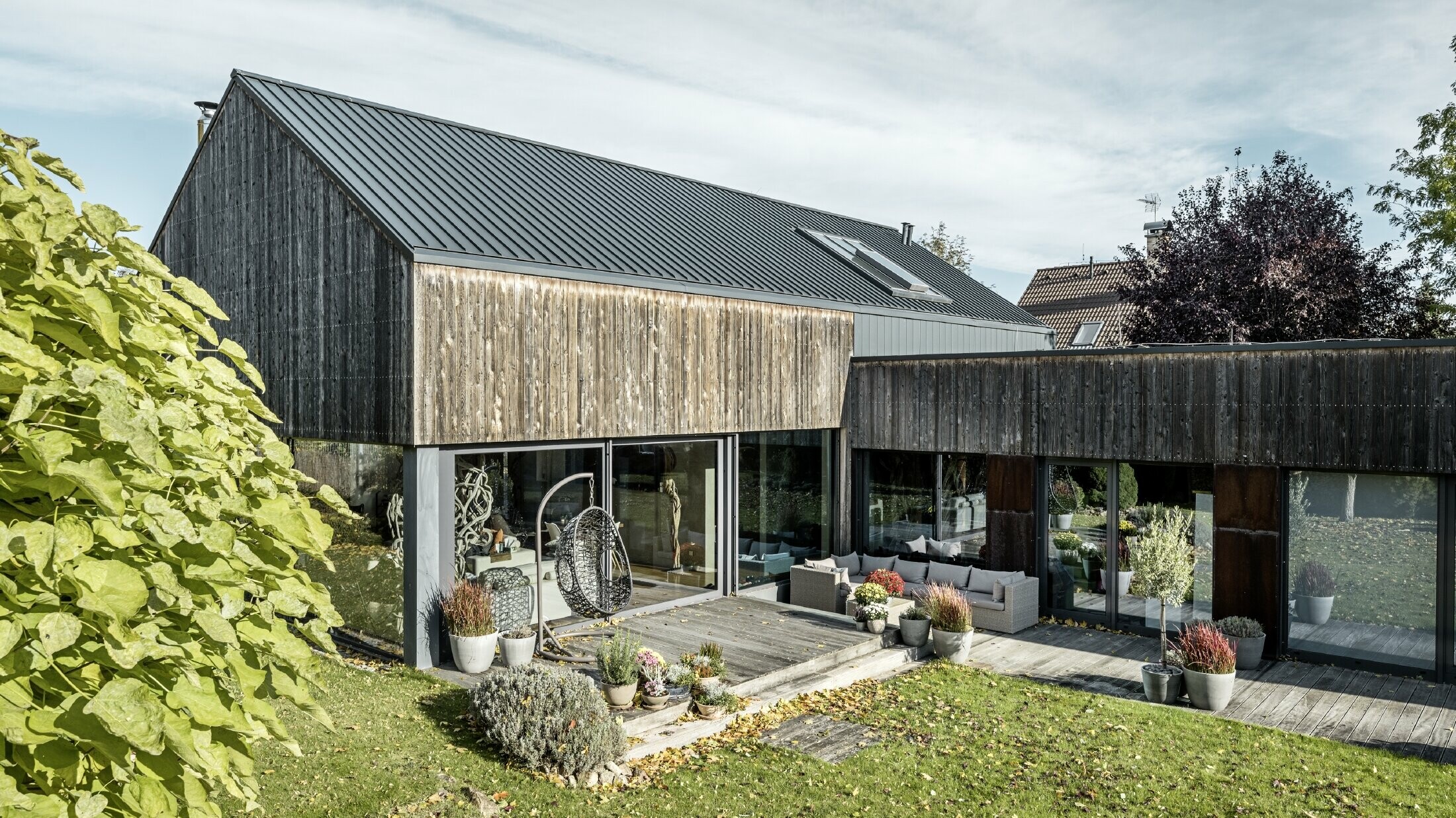 Dom jednorodzinny z dachem dwuspadowym, pokryty PREFALZ w technologii rąbka podwójnego w kolorze antracytowym z elewacją z postarzanego drewna. Z pięknym tarasem drewnianym i dużymi oknami na parterze.
