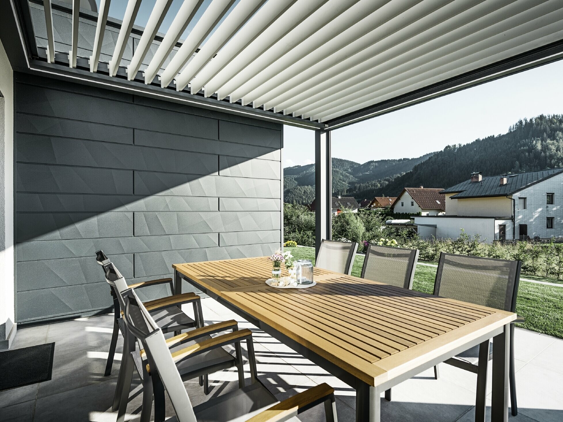 Zadaszony taras z lamelami, wyposażony w stół i krzesła; jedna ze ścian elewacji wykończona panelami PREFA Siding.X w kolorze antracyt.