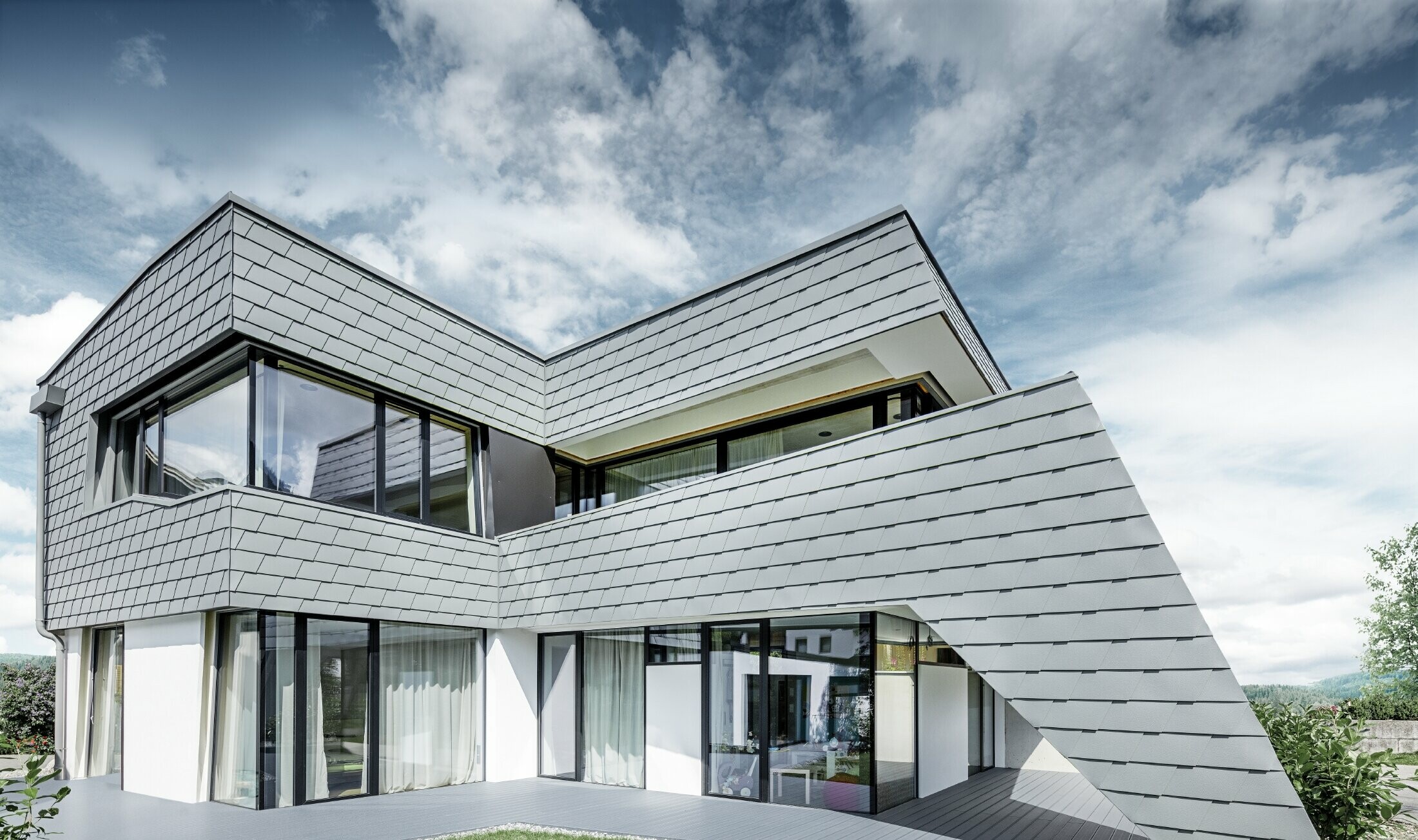 Nowoczesny dom jednorodzinny z płaskim dachem, dużymi powierzchniami okiennymi i aluminiowym łupkiem fasadowym PREFA w kolorze jasnoszarym