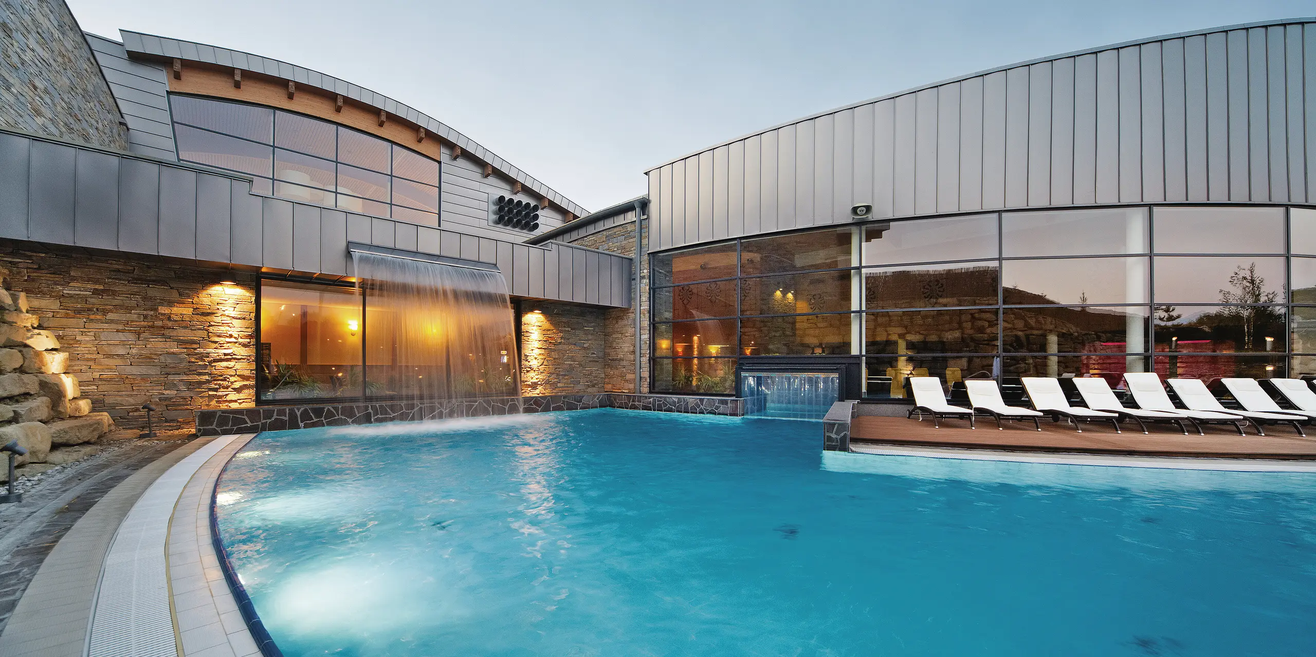 In der Dämmerung wird die Therme Białka durch die beleuchteten Fenster und die reflektierenden Wasserflächen besonders in Szene gesetzt. Die PREFALZ-Fassade in der Sonderfarbe Delphin unterstreicht die elegante Architektur, die durch die Kombination von Aluminium und Naturmaterialien eine harmonische und zeitlose Wirkung erzielt.
