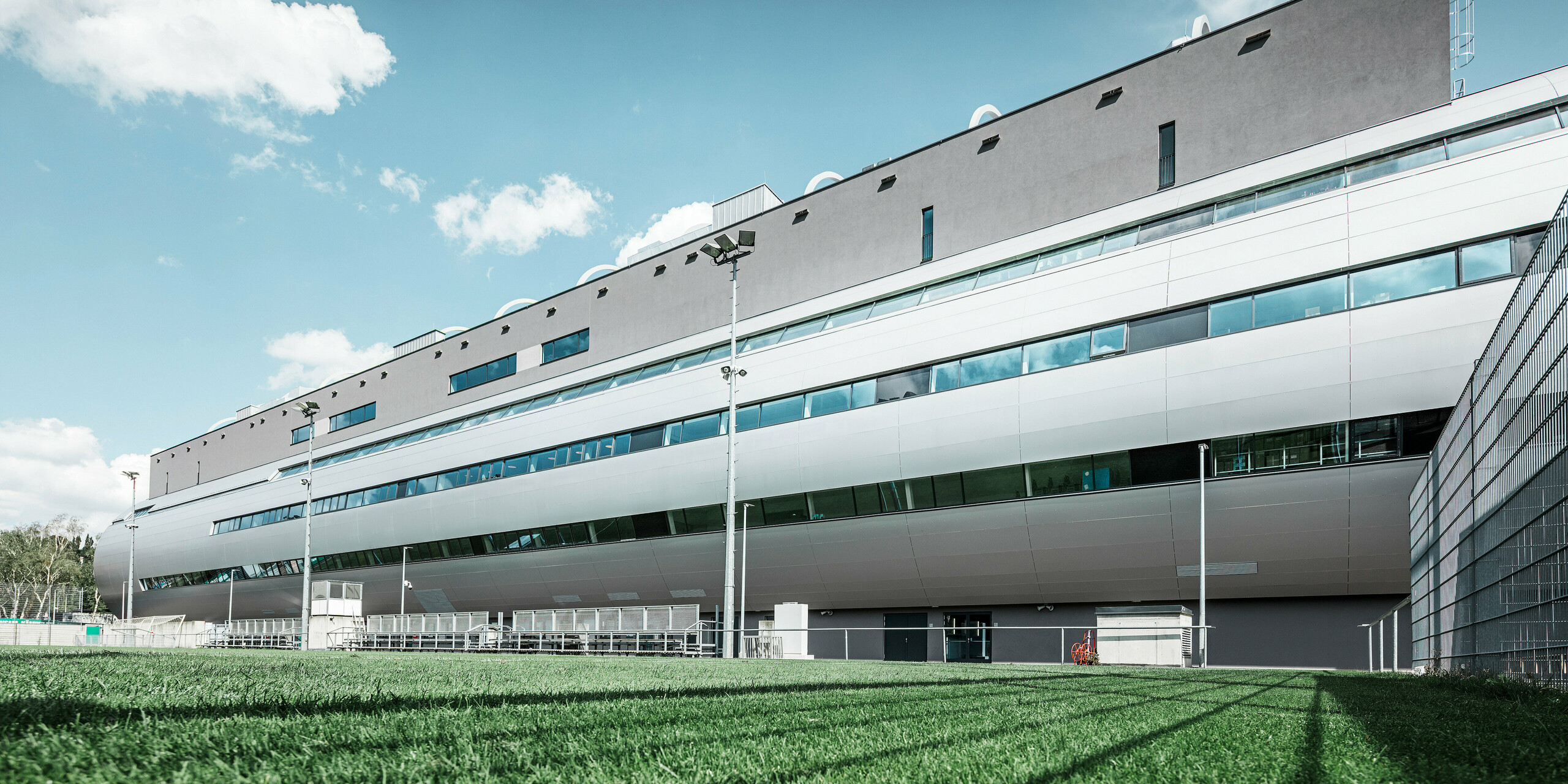Das Allianz Stadion von SK Rapid Wien, abgebildet vom Trainingsplatz, zeigt die glänzende Fassade aus PREFA Aluminium Verbundplatten in Silbermetallic. Die harmonische Verschmelzung von funktionaler Sportstätte und eleganter Architektur wird durch die Fensterreihen betont. Die grüne Wiese des Trainingsplatzes am unteren Bildrand wird durch die Betreuerbänke und den Zaun abgegrenzt. Am Zaus sind zudem hohe Flutlichtmasten zu erkennen, die eine Ausübung des Trainings zu jeder Tageszeit ermöglichen. Der blaue Himmel komplementiert das professionelle Foto.