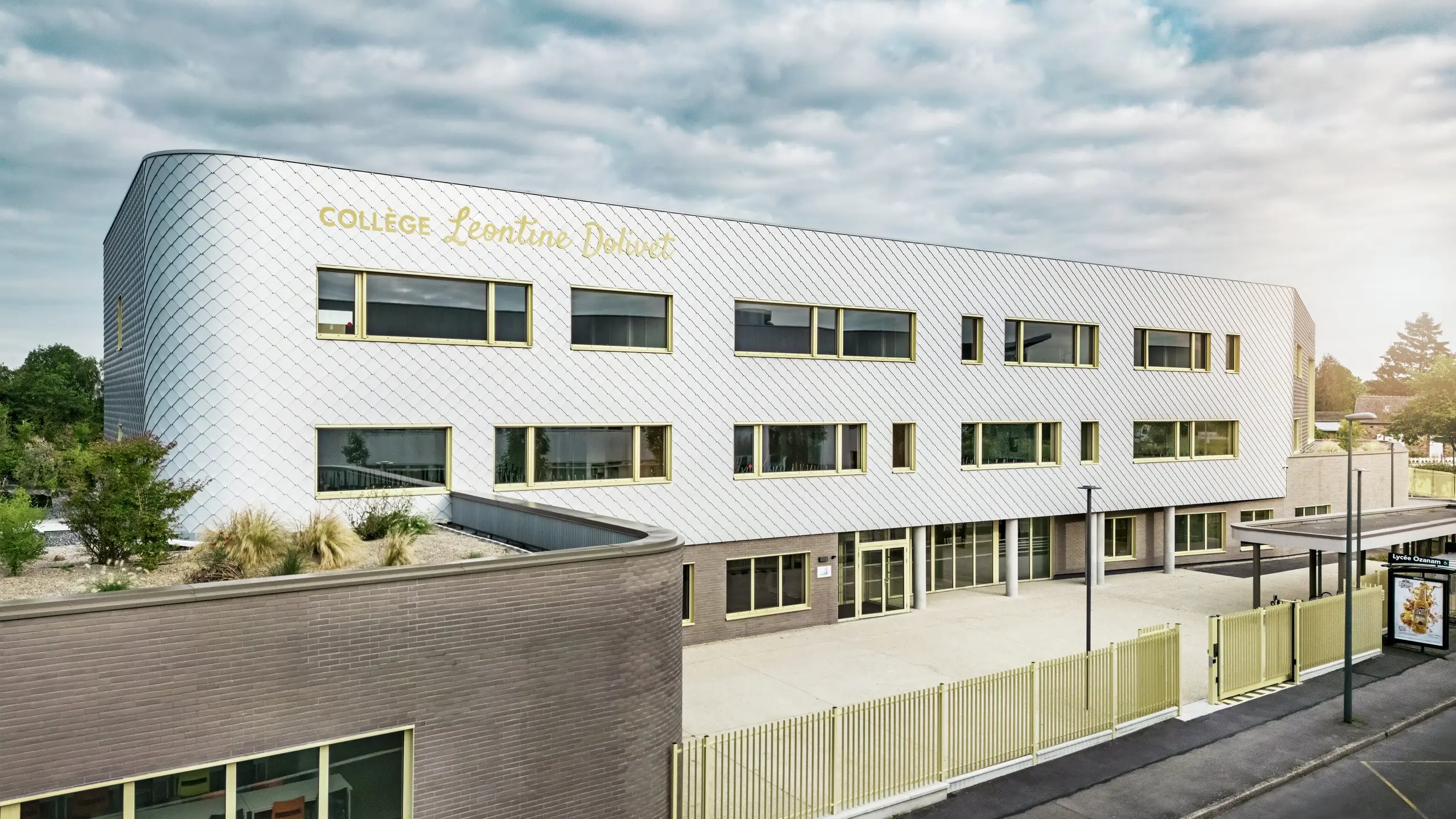 Die Vorderansicht des Collège Léontine Dolivet in Cesson-Sévigné zeigt eine moderne Aluminiumfassade aus PREFA-Wandelementen in Silbermetallic. Die variable Oberfläche verleiht dem Gebäude Leichtigkeit, während klare Linien und große Fenster für Transparenz sorgen. Gelbe Fensterrahmen setzen farbliche Akzente. Im Vordergrund sind der helle Eingangsbereich, der Zaun und die angrenzende Straße zu sehen. Die langlebige PREFA-Fassadenlösung vereint Design, Witterungsbeständigkeit und Nachhaltigkeit – ein architektonisch ansprechendes Gebäude mit einer eleganten Formensprache.