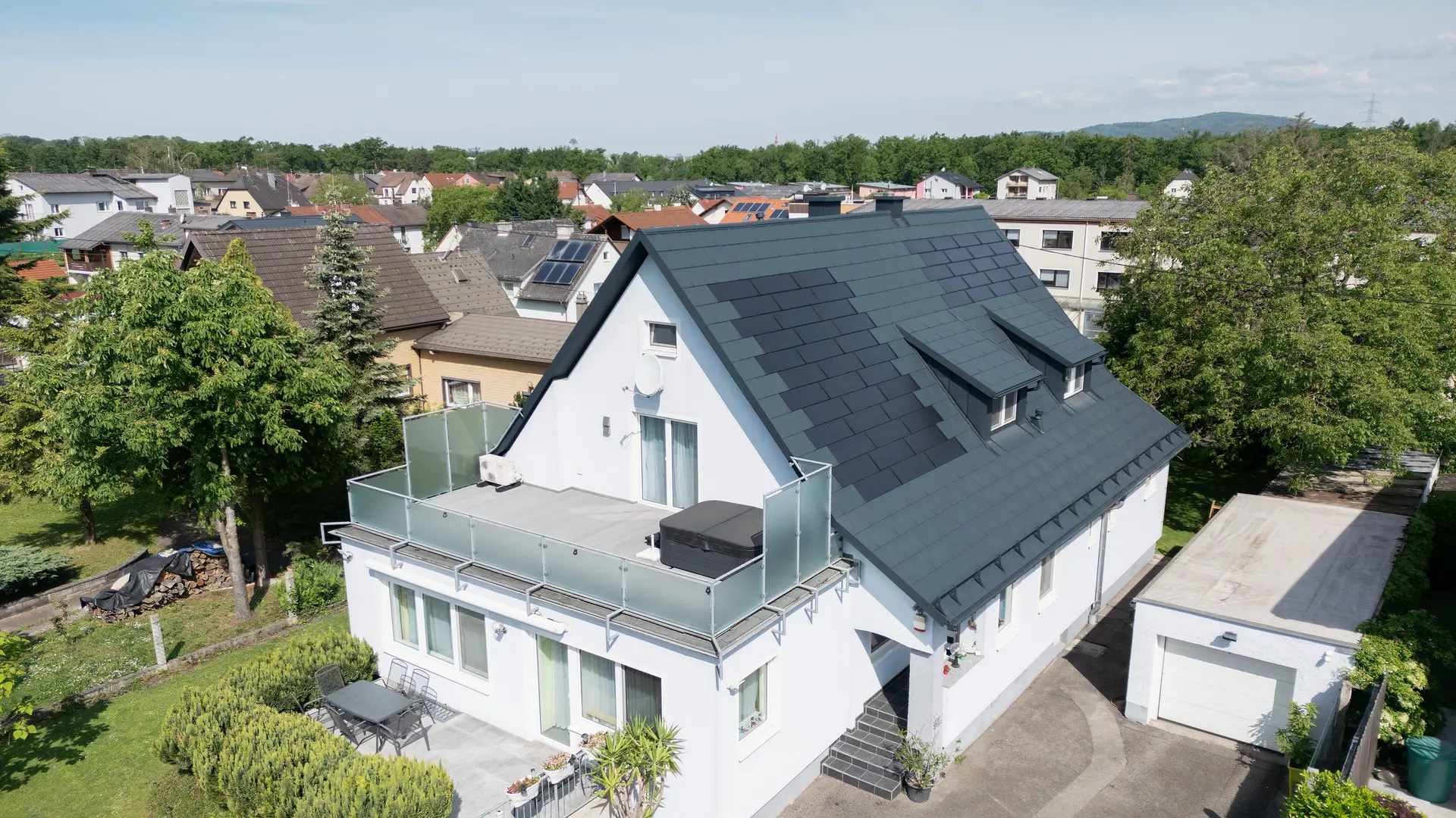 Widok z lotu ptaka na dom jednorodzinny po renowacji dachu. Dach pokryty jest teraz dużymi dachówkami solarnymi PREFA oraz dachówką PREFA R.16 w kolorze P.10 antracyt. Dachówki solarne są rozmieszczone równomiernie, tworząc nowoczesne i ekologiczne rozwiązanie. Budynek prezentuje się teraz nowocześnie i przyjaznie dla środowiska. Balkon na pierwszym planie ma szklaną balustradę i znajduje się na najwyższym piętrze budynku.