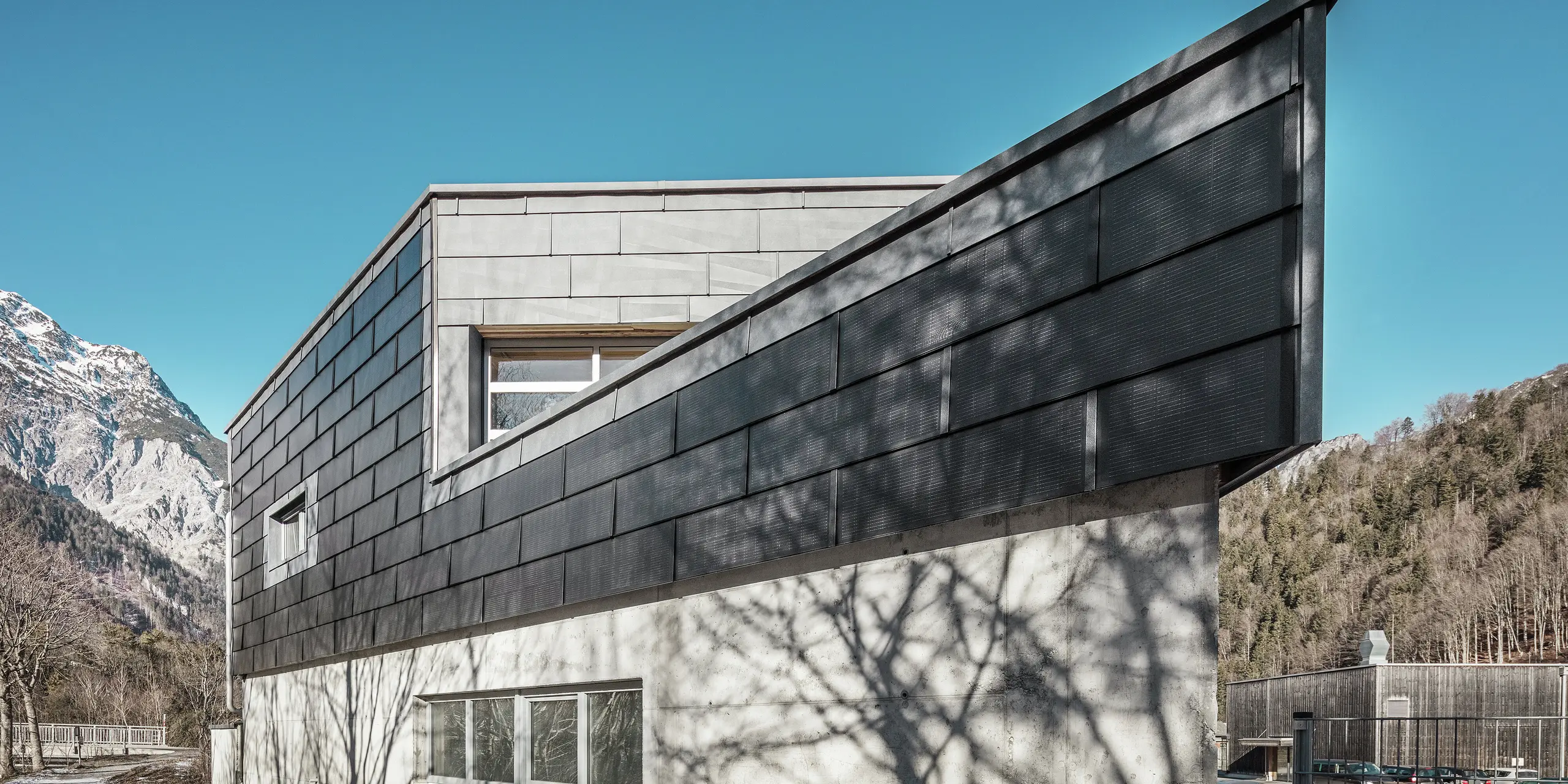 Das Bürogebäude der RÜMA Blechwerkstatt in Vorarlberg besticht durch seine moderne Fassade aus PREFA Solardachpaneelen und Sichtbeton. Umgeben von Bergen und Bäumen, deren Schatten sich an der Wand abzeichnen, erhebt es sich unter strahlend blauem Himmel. Die markante Gebäudekante und die PREFA Fassadenpaneele FX.12 unterstreichen den eindrucksvollen Eindruck zusätzlich.