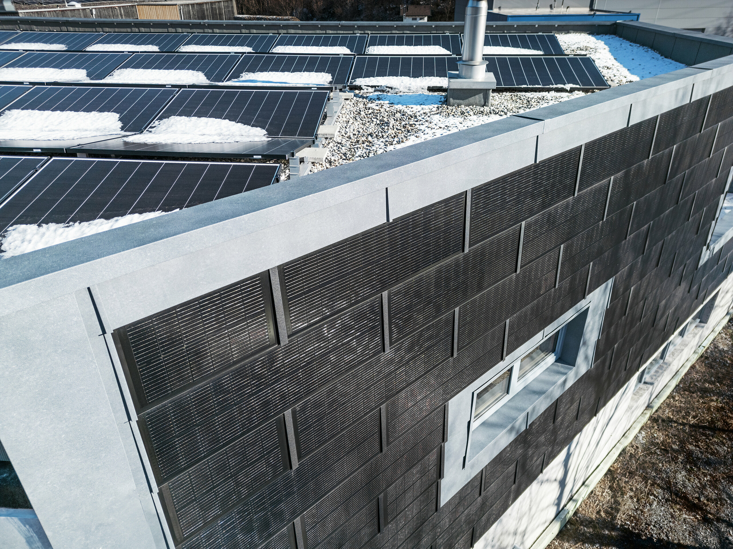Die Luftaufnahme zeigt die Fassade der RÜMA-Blechwerkstatt in Vorarlberg mit grauen FX.12-Fassadenpaneelen und innovativen PREFA-Solardachpaneelen mit integrierten Photovoltaikmodulen. Im Vordergrund ist ein teilweise schneebedecktes Flachdach mit weiteren Photovoltaikmodulen zu sehen.