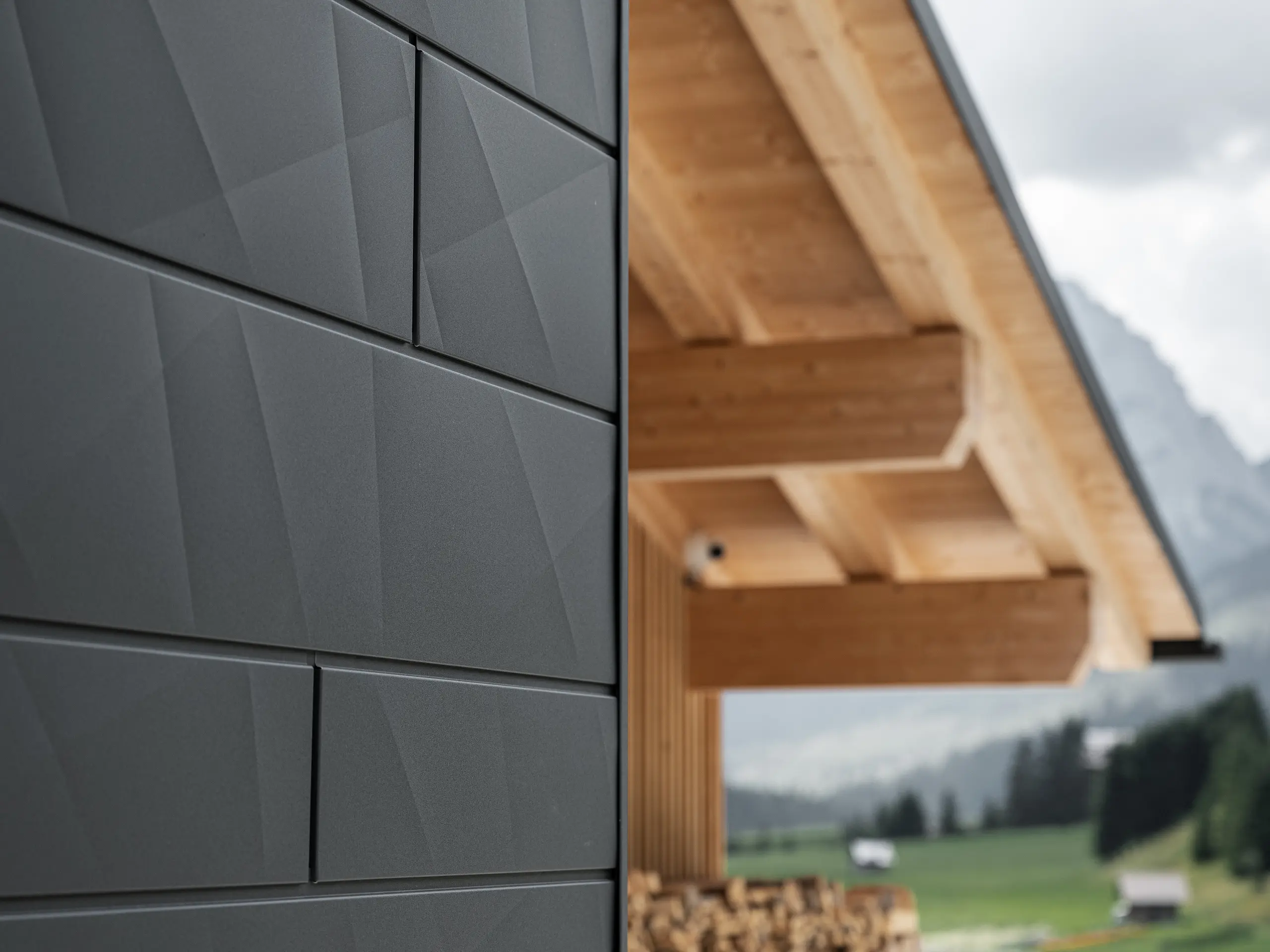 Zbliżenie paneli elewacyjnych Siding.X w kolorze P.10 czarny od PREFA na budynku Casa Hoffe w Sappadzie we Włoszech. Aluminiowy system elewacyjny został umiejętnie zastosowany w nowoczesnym, abstrakcyjnym układzie, który tworzy atrakcyjny kontrast z widocznymi drewnianymi elementami więźby dachowej. Innowacyjne rozwiązanie elewacyjne harmonijnie wpisuje się w alpejskie otoczenie i symbolizuje połączenie funkcjonalności, trwałości oraz estetycznej kultury budowlanej.