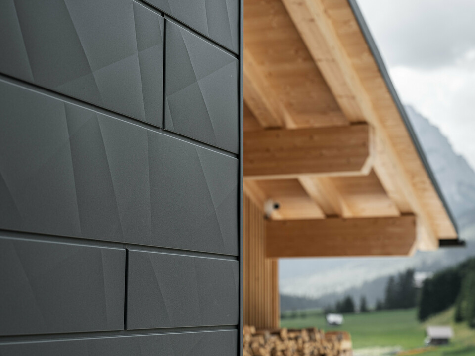 Zbliżenie paneli elewacyjnych Siding.X w kolorze P.10 czarnym firmy PREFA na budynku Casa Hoffe w Sappadzie we Włoszech. System elewacyjny z aluminium został umiejętnie zastosowany w nowoczesnym, abstrakcyjnym projekcie, który stanowi atrakcyjny kontrast dla widocznych elementów drewnianych konstrukcji dachu. Innowacyjne rozwiązanie elewacyjne idealnie komponuje się z alpejskim otoczeniem i stanowi połączenie funkcjonalności, trwałości i estetycznej kultury budowlanej.