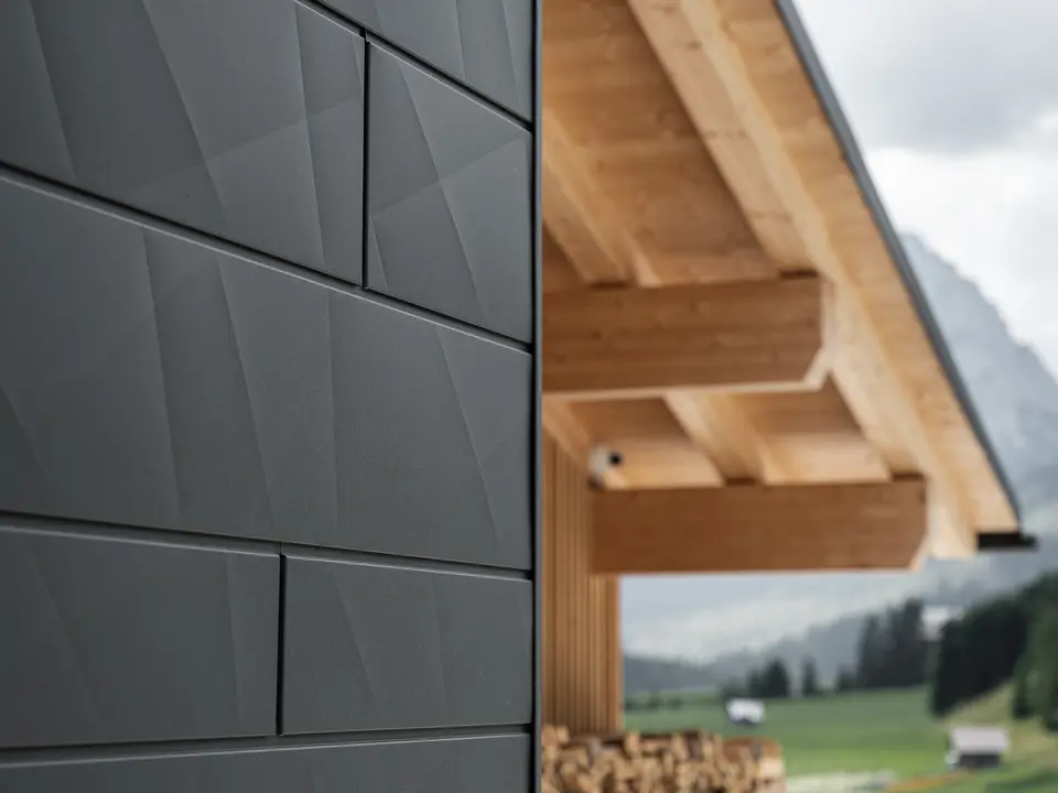 Zbliżenie paneli elewacyjnych Siding.X w kolorze P.10 czarny od PREFA na budynku Casa Hoffe w Sappadzie we Włoszech. Aluminiowy system elewacyjny został umiejętnie zastosowany w nowoczesnym, abstrakcyjnym układzie, który tworzy atrakcyjny kontrast z widocznymi drewnianymi elementami więźby dachowej. Innowacyjne rozwiązanie elewacyjne harmonijnie wpisuje się w alpejskie otoczenie i symbolizuje połączenie funkcjonalności, trwałości oraz estetycznej kultury budowlanej.