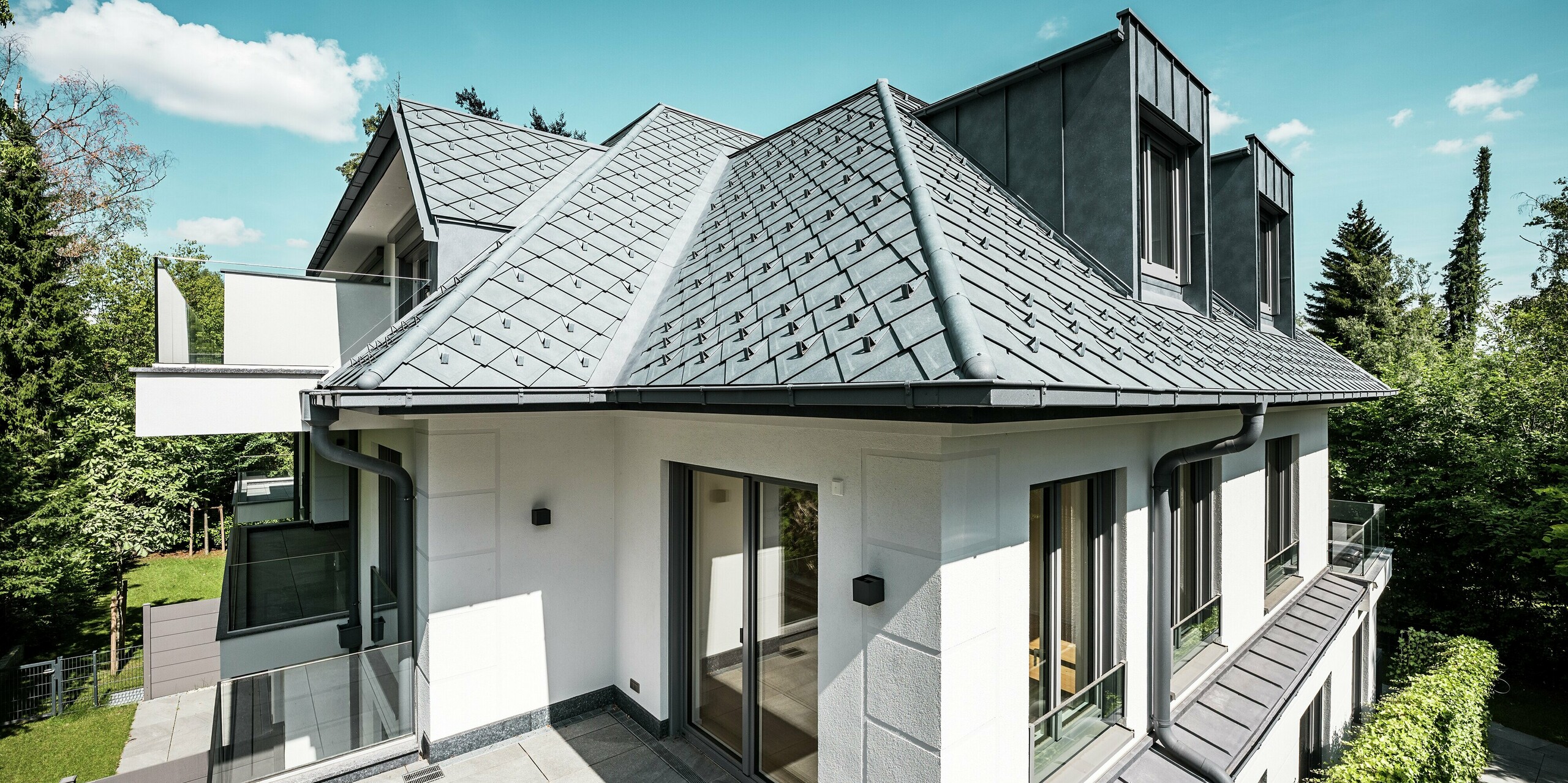 Modernes Mehrfamilienhaus mit markanter Dacharchitektur in PREFA P.10 Steingrau: Die Dachfläche ist mit hochwertigen PREFA Dachrauten 29 × 29 gedeckt und sorgt für ein edles, strukturiertes Erscheinungsbild. Stilvolle Flachdachgauben aus PREFALZ fügen sich nahtlos ins Design ein. Integrierte Schneestopper und eine formschöne PREFA Dachentwässerung mit Kastenrinne und Ablaufrohr unterstreichen die technische Raffinesse. Das langlebige Aluminiumdach trotzt jeder Witterung und vereint Ästhetik mit Funktionalität – ideal für zeitgemäßen Wohnbau.