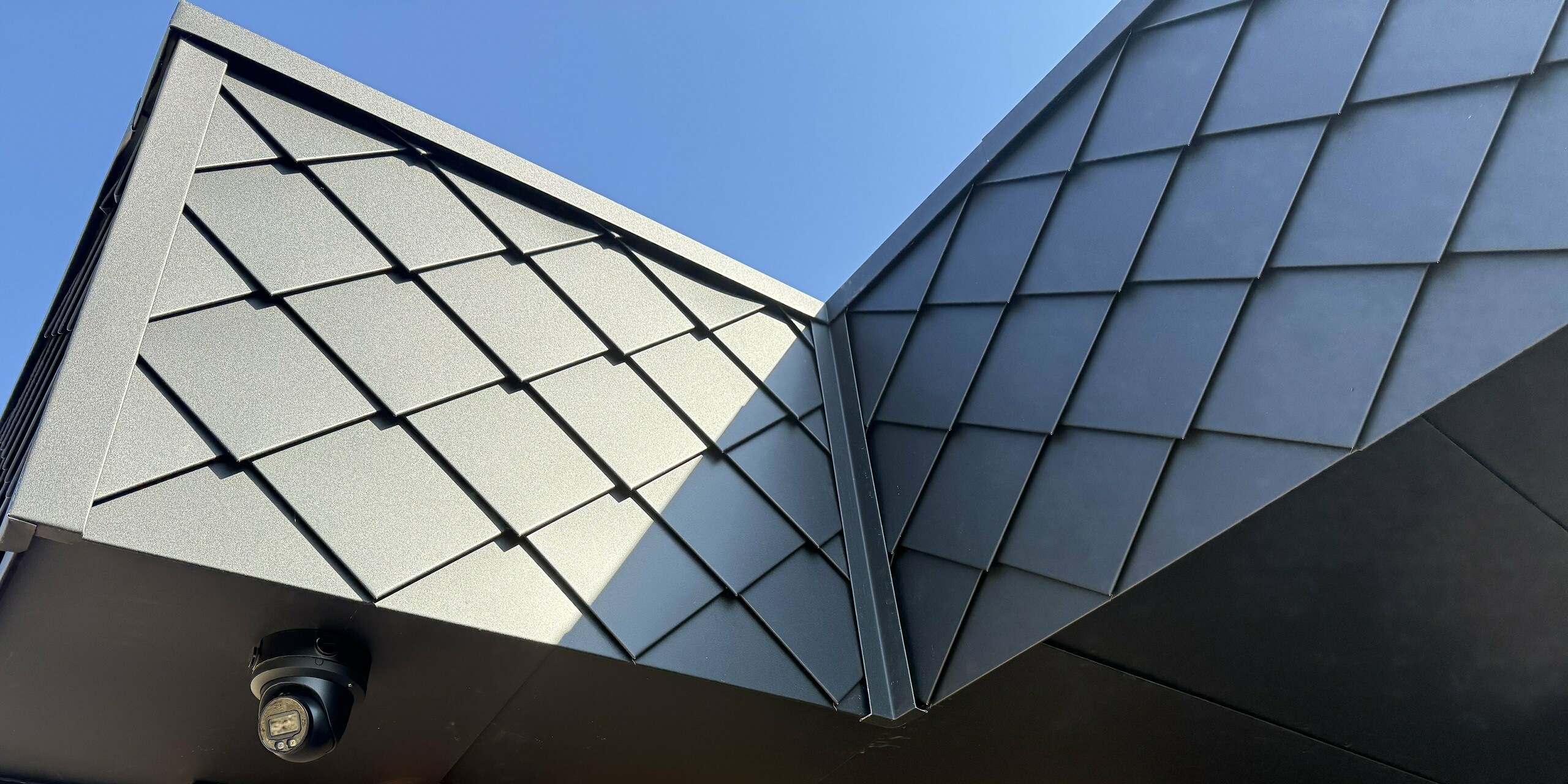 Detailansicht einer Gebäudeecke mit markanter Fassadengestaltung: Die kleinformatige PREFA Wandraute 20 × 20 in P.10 Anthrazit setzt elegante Akzente und betont die dynamische Linienführung. Im Kontrast zur warmen Klinkerfassade entsteht ein spannender Materialmix, der modernes Design und höchste Qualität vereint. Ideal für nachhaltige Architektur mit unverwechselbarem Charakter.