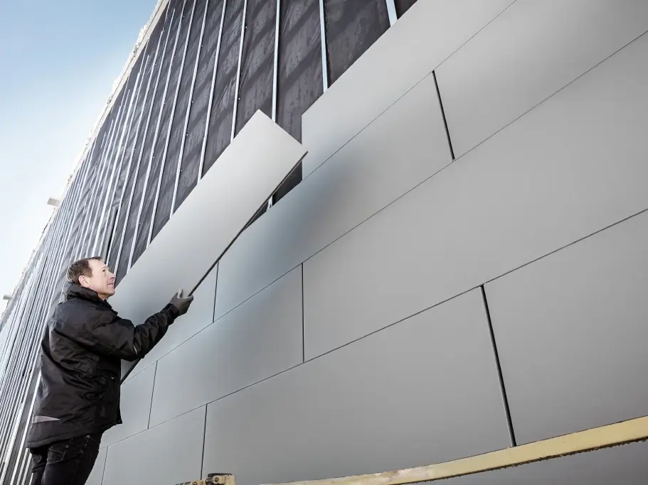 Zdjęcia częściowo ukończonej elewacji wykończonej sidingiem PREFA o szerokości 500. Dekarz układa kolejny panel elewacyjny. Zdjęcie zrobione z dołu. 