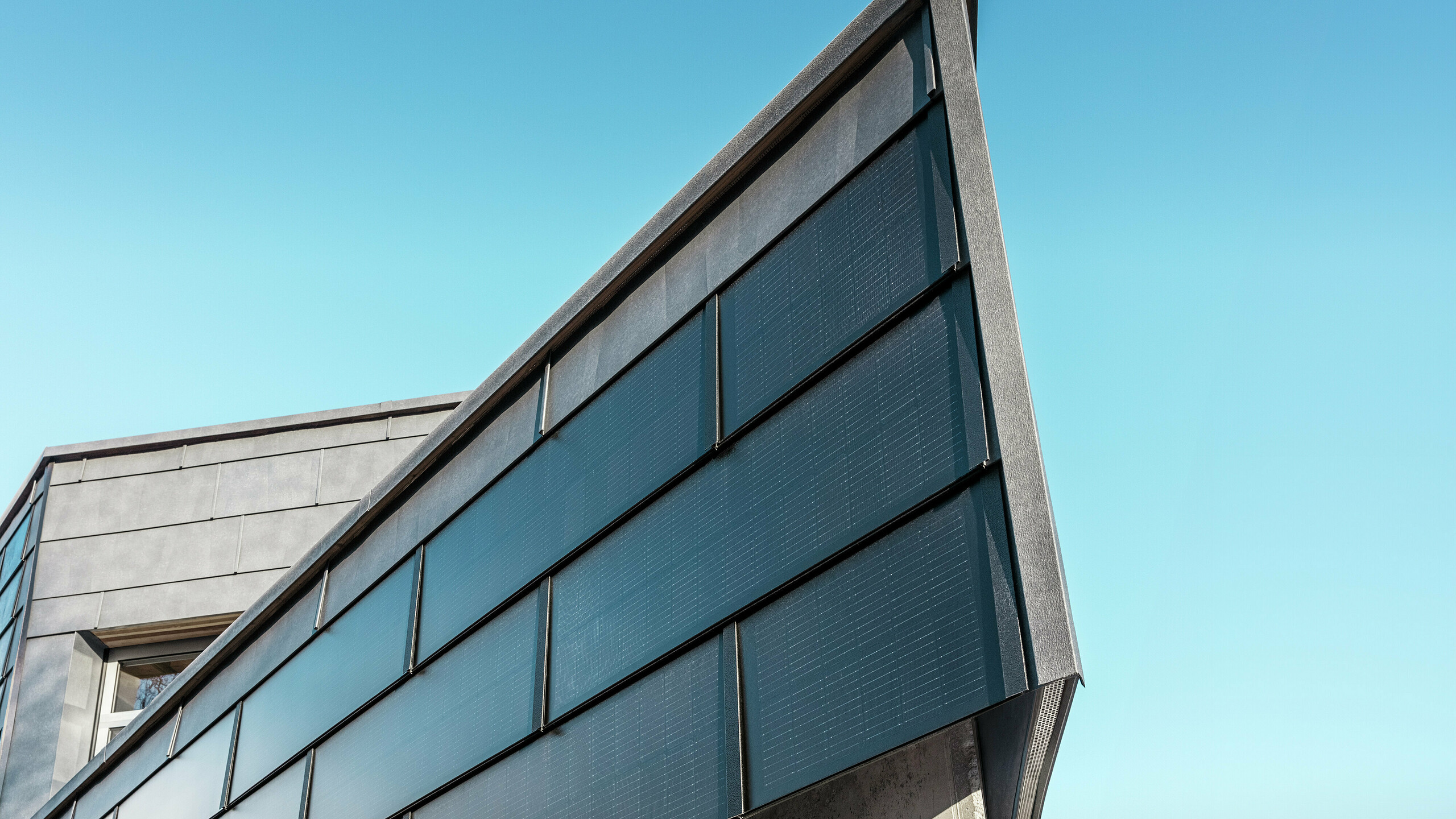 Moderne Geschäftsfassade der RÜMA Blechwerkstatt in Vorarlberg mit PREFA Solar-Aluminium-Dachpaneelen und integrierten Photovoltaikmodulen, kombiniert mit PREFA Fassadenpaneelen FX.12 für nachhaltige Energieerzeugung und dynamische Fassadengestaltung. Die Aufnahme zeigt eine Nahaufnahme einer Gebäudeecke. Im Hintergrund ist ein klarer blauer Himmel zu sehen.