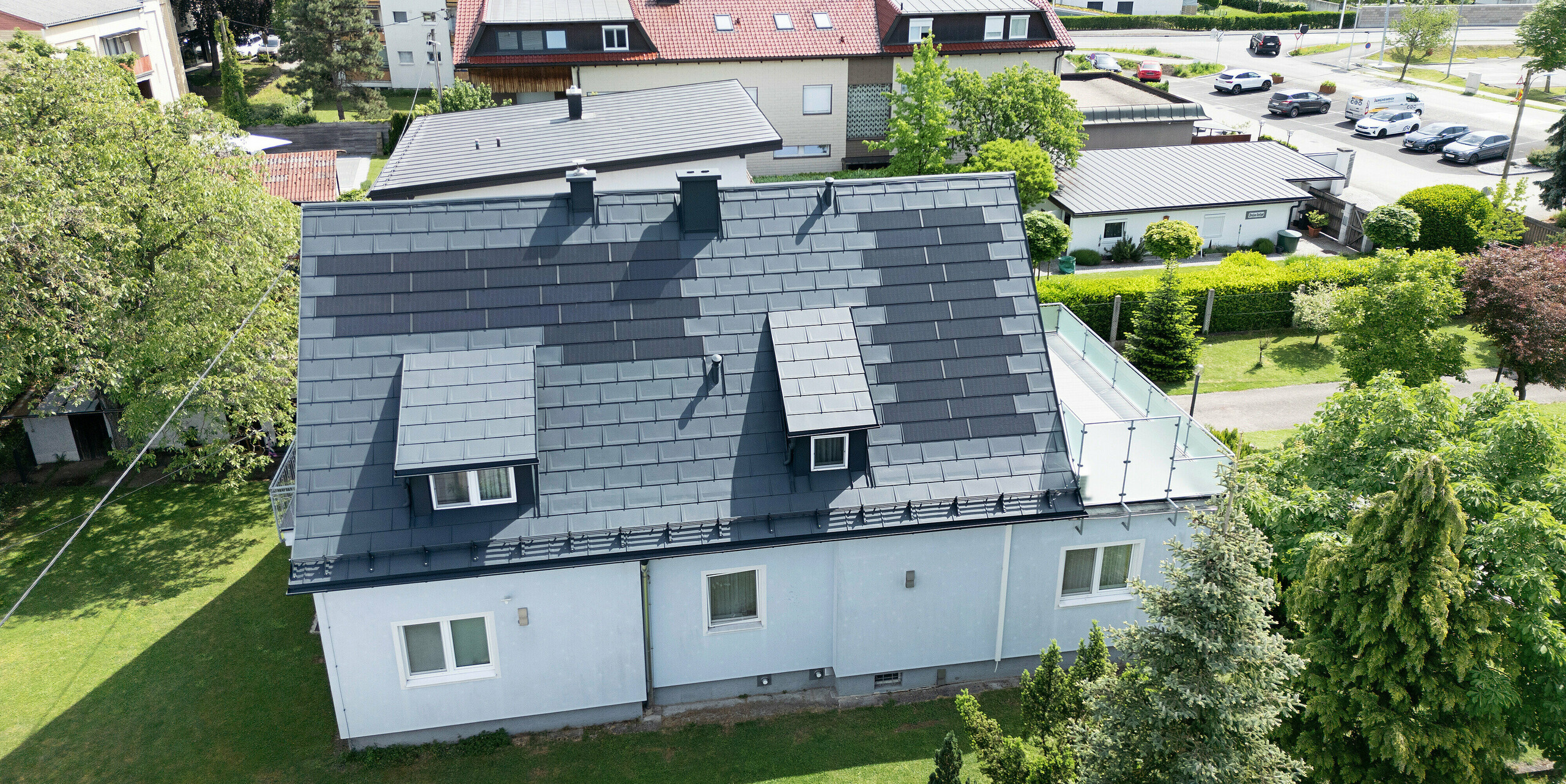 Saniertes Dach eines Einfamilienhauses in Leonding mit PREFA Solardachplatten in Anthrazit. Die Vogelperspektive zeigt das modernisierte PREFA Dach aus hochwertigem Blech mit integrierten Solarmodulen. Diese langlebige und witterungsbeständige Lösung bietet eine nachhaltige und energieeffiziente Möglichkeit zur Stromerzeugung. Das Bild verdeutlicht die ästhetische Integration der Solarmodule und die funktionale Gestaltung der traditionellen Schleppgauben. Die Flächen ohne PV-Module wurden mit der robusten PREFA Dachplatte R.16 in der Farbe P.10 Anthrazit verkleidet. 