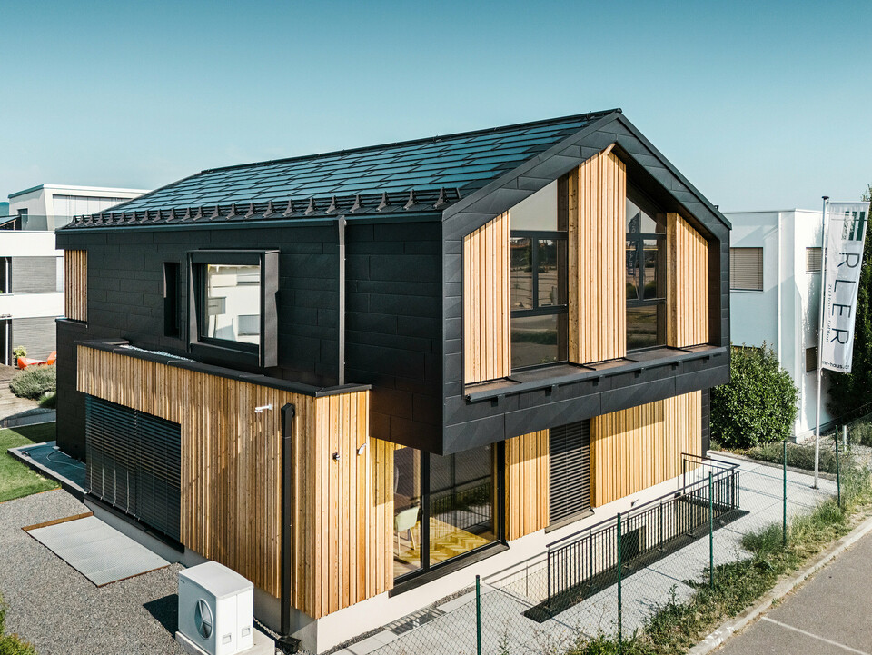 Przód domu pokazowego Autark w centrum domów prefabrykowanych Blaue Lagune, Vösendorf (AT). Dach pokryty jest czarnymi dachówkami solarnymi PREFA i dachówkami R.16 z wysokiej jakości powłoką P.10. Elewacja składa się z połączenia pionowych elementów drewnianych i PREFA Siding.X w P.10 czarnym, co nadaje budynkowi nowoczesny i ekologiczny charakter. Na prawym brzegu zdjęcia powiewa flaga firmy Erler Bau GmbH.