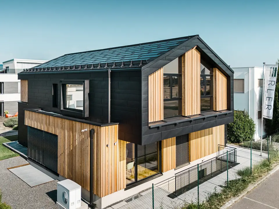 Przód domu pokazowego Autark w centrum domów prefabrykowanych Blaue Lagune, Vösendorf (AT). Dach pokryty jest czarnymi dachówkami solarnymi PREFA i dachówkami R.16 z wysokiej jakości powłoką P.10. Elewacja składa się z połączenia pionowych elementów drewnianych i PREFA Siding.X w P.10 czarnym, co nadaje budynkowi nowoczesny i ekologiczny charakter. Na prawym brzegu zdjęcia powiewa flaga firmy Erler Bau GmbH.