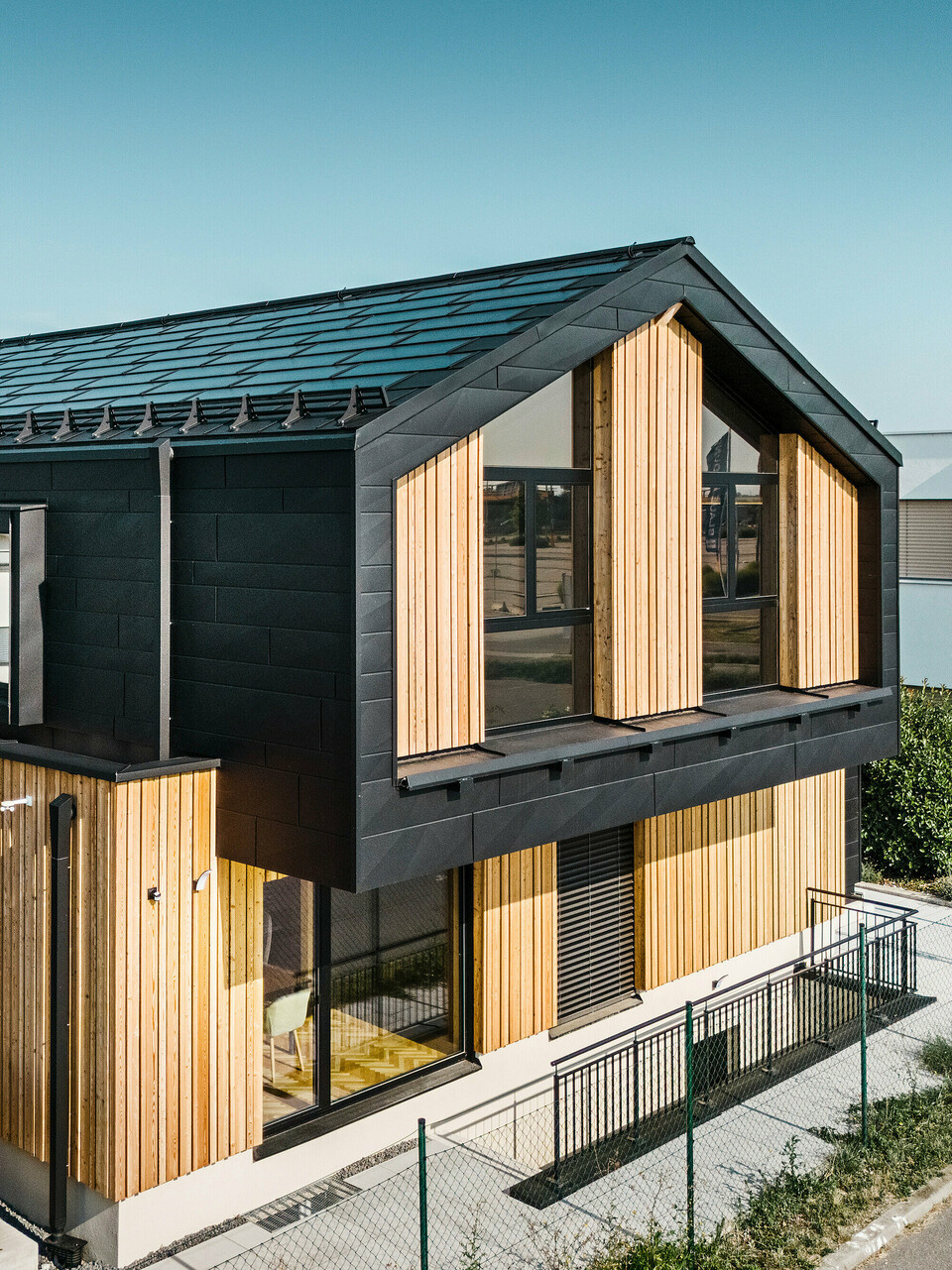 Przód domu pokazowego Autark w centrum domów prefabrykowanych Blaue Lagune, Vösendorf (AT). Dach pokryty jest czarnymi dachówkami solarnymi PREFA i dachówkami R.16 z wysokiej jakości powłoką P.10. Elewacja składa się z połączenia pionowych elementów drewnianych i PREFA Siding.X w P.10 czarnym, co nadaje budynkowi nowoczesny i ekologiczny charakter. Na prawym brzegu zdjęcia powiewa flaga firmy Erler Bau GmbH.
