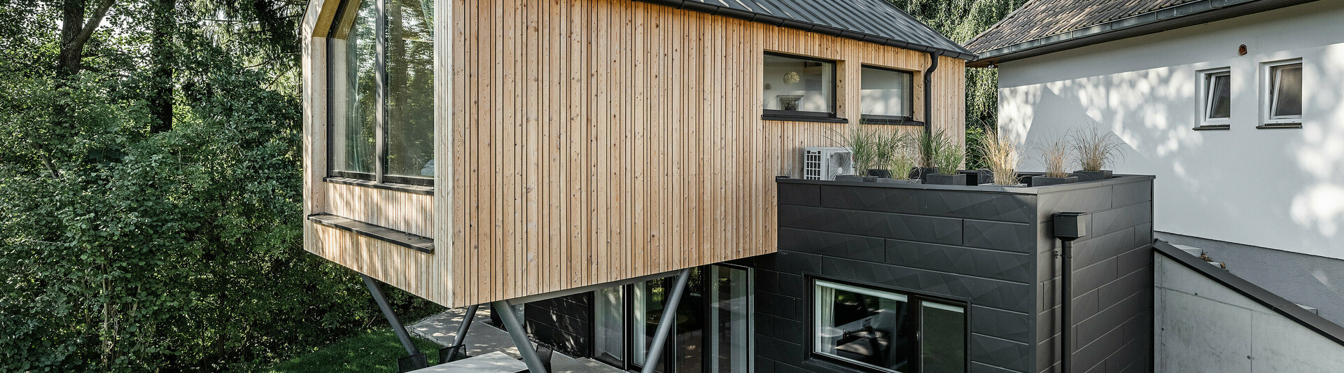 Das 'Woodstock Chalet 5' beeindruckt mit einer eindrucksvollen Kombination aus natürlichen Holzoberflächen und den modernen PREFA Siding.X Fassadenelementen in Anthrazit. Die überhängende Holzstruktur des Obergeschosses ragt markant über die dunklen Fassadenelemente, die das Erdgeschoss verkleiden, und erzeugt so ein Spiel aus Licht und Schatten. Die klaren Linien und die robuste Konstruktion der Siding.X Elemente ergänzen das innovative Design des Gebäudes und bieten einen spannenden visuellen Kontrast zur umgebenden Vegetation und dem traditionellen Gebäude im Hintergrund. Am Dach des Chalets wurde das PREFALZ Dachsystem in Form einer gleichmäßigen Stehfalzdeckung verlegt. Am rechten Rand des Erdgeschosses ist das PREFA Dachentwässerungssystem zu erkennen, das analog zur Aluminiumfassade in Anthrazit gehalten ist. Für die Dachentwässerung wurden der Wasserfangkasten gefolgt vom Ablaufrohr montiert.