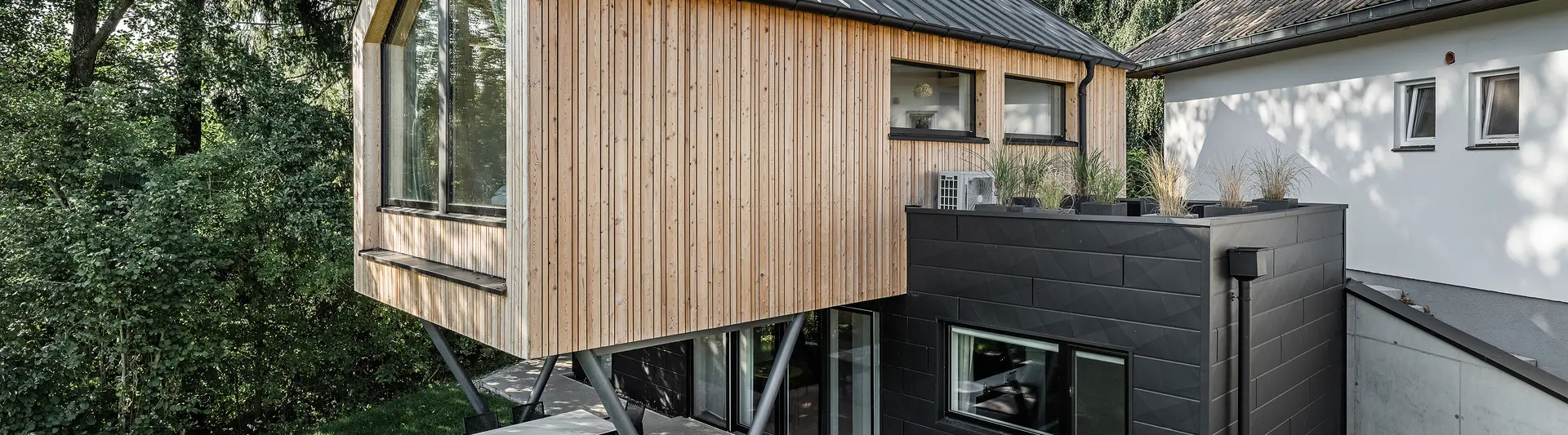 Das 'Woodstock Chalet 5' beeindruckt mit einer eindrucksvollen Kombination aus natürlichen Holzoberflächen und den modernen PREFA Siding.X Fassadenelementen in Anthrazit. Die überhängende Holzstruktur des Obergeschosses ragt markant über die dunklen Fassadenelemente, die das Erdgeschoss verkleiden, und erzeugt so ein Spiel aus Licht und Schatten. Die klaren Linien und die robuste Konstruktion der Siding.X Elemente ergänzen das innovative Design des Gebäudes und bieten einen spannenden visuellen Kontrast zur umgebenden Vegetation und dem traditionellen Gebäude im Hintergrund. Am Dach des Chalets wurde das PREFALZ Dachsystem in Form einer gleichmäßigen Stehfalzdeckung verlegt. Am rechten Rand des Erdgeschosses ist das PREFA Dachentwässerungssystem zu erkennen, das analog zur Aluminiumfassade in Anthrazit gehalten ist. Für die Dachentwässerung wurden der Wasserfangkasten gefolgt vom Ablaufrohr montiert.
