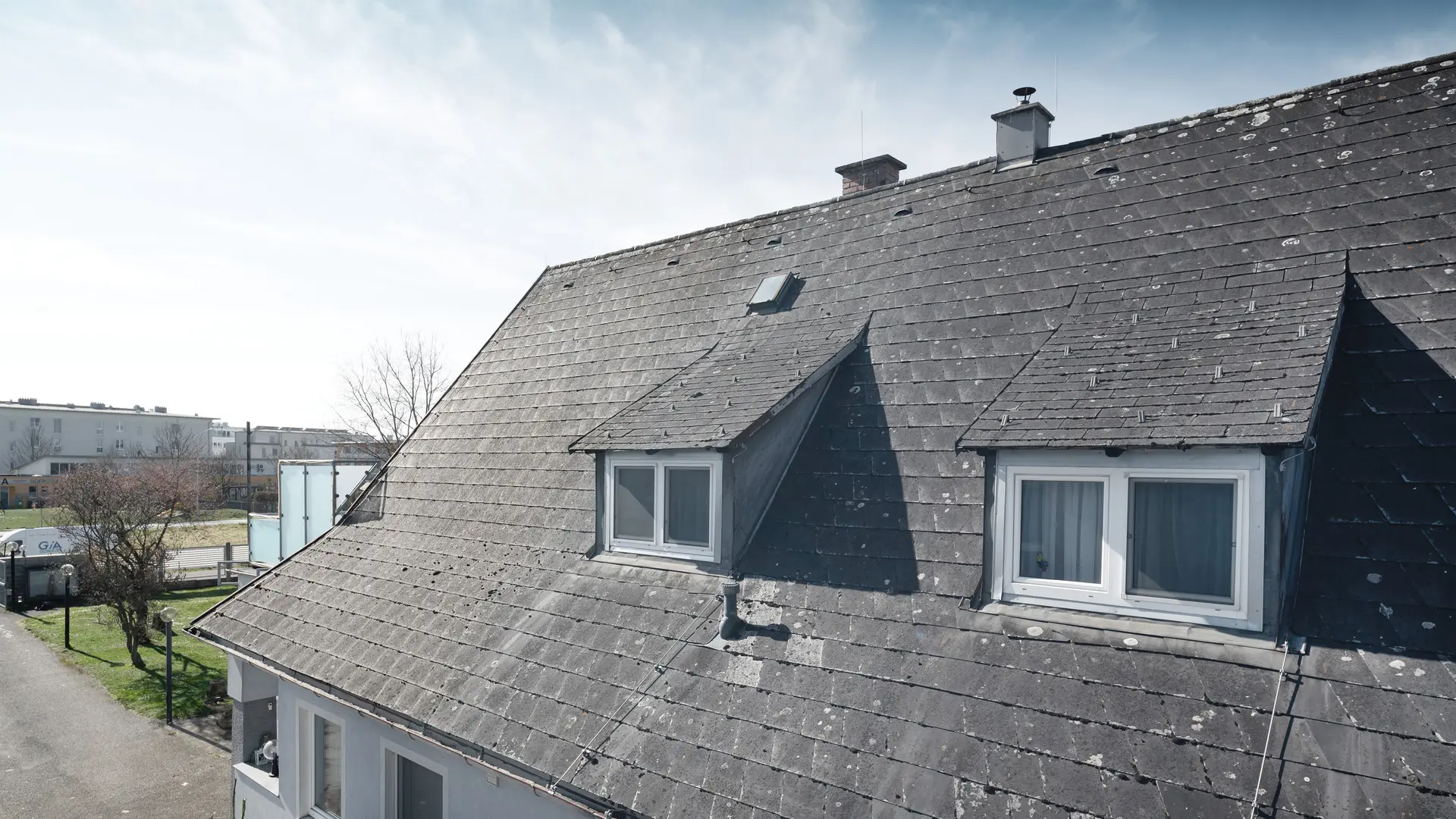 Zbliżenie starego dachu z płyt Eternit z widocznymi śladami zużycia i porostami. Dach posiada dwa widoczne lukarny. Zdjęcie wykonane przed renowacją dachu.