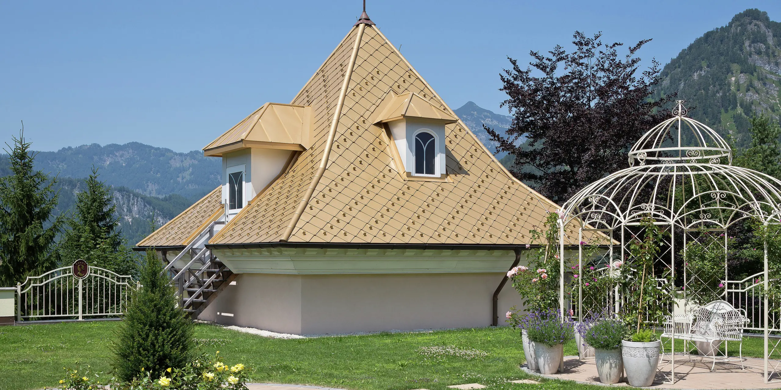 Kleiner Zubau eines Einfamilienhauses in Tirol mit PREFA Dachrauten in Mayagold und einem malerischen Garten im Vordergrund, umgeben von den Alpen.
