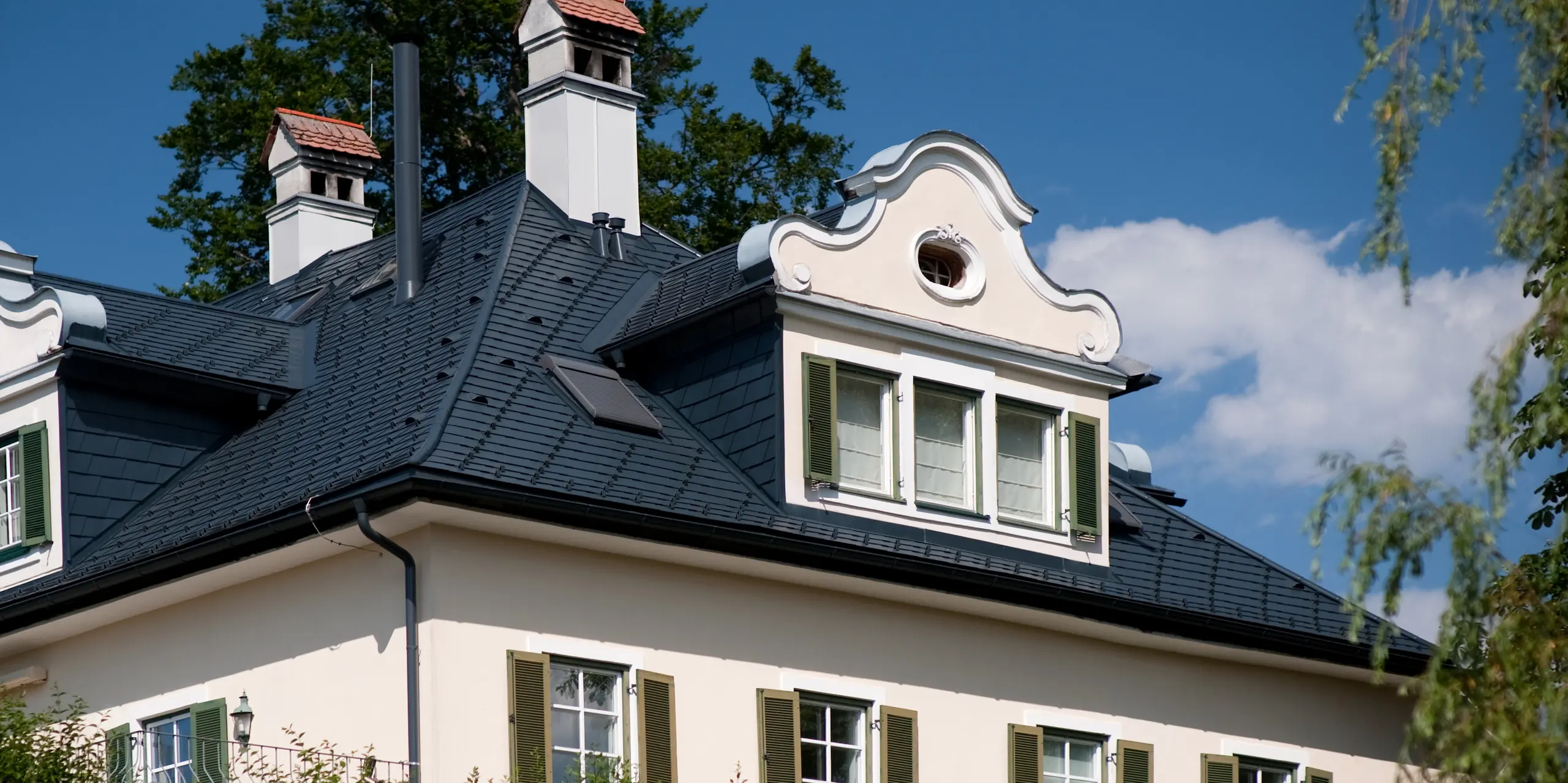 Detailansicht der denkmalgeschützten Villa in Bad Aussee mit markantem Dach aus PREFA Dachschindeln in P.10 Anthrazit. Die elegante Dachlandschaft mit Gauben, Kaminen und geschwungenem Giebel setzt architektonische Akzente. Die langlebigen Aluminium-Dachschindeln überzeugen durch ihre hochwertige Optik, Wetterbeständigkeit und den zuverlässigen Schutz für Generationen.