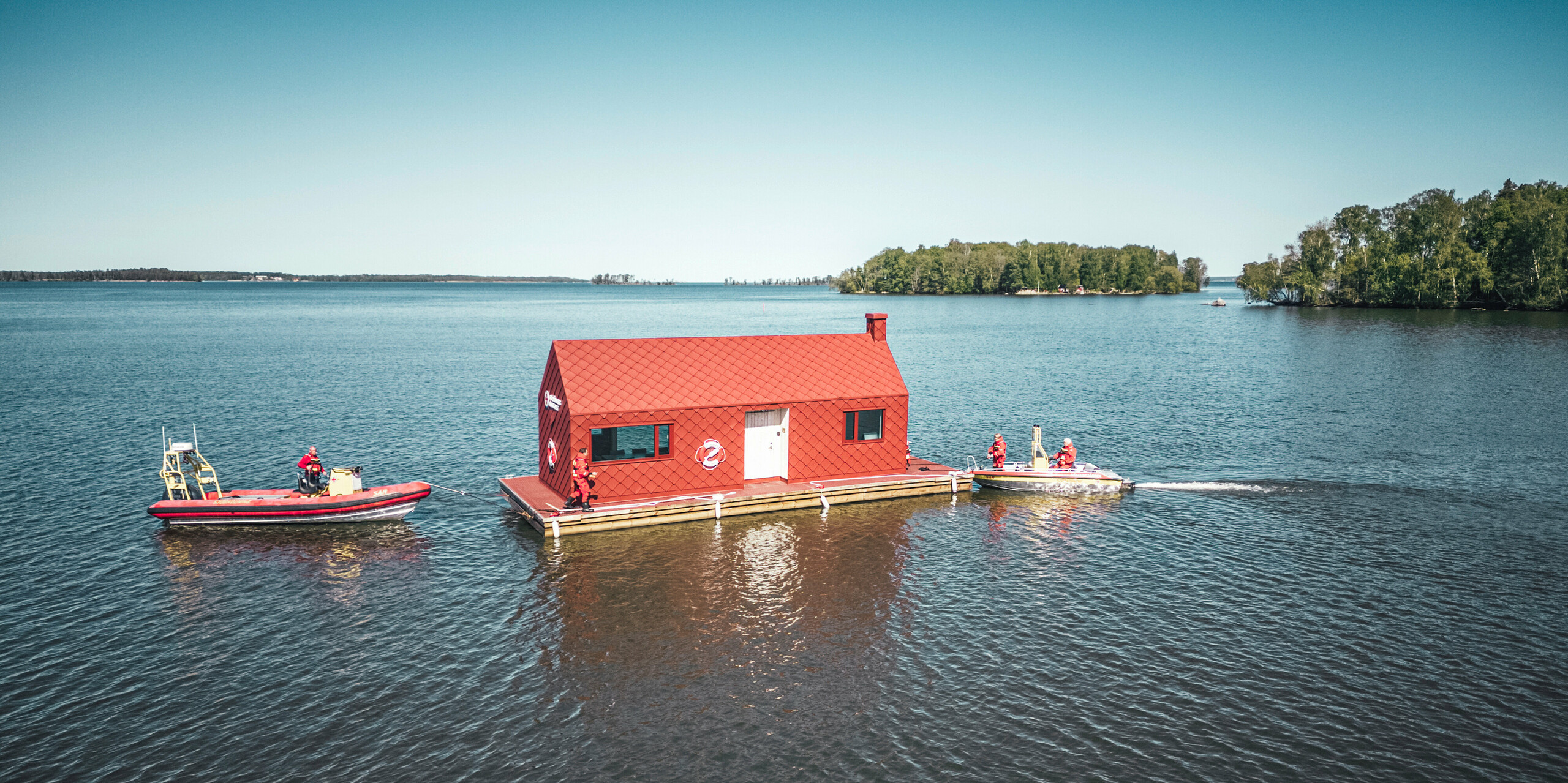 [Translate to Polski:] Luftaufnahme des Seenotrettungsstationshauses auf dem Hjälmaren See in Schweden, umgeben von der ruhigen Weite des Wassers. Das markante, oxydrote Gebäude, ausgestattet mit PREFA Dach- und Wandrauten 29×29, thront auf einem Schwimmkörper und ist bereit, den Rettungskräften Zuflucht und Betriebsbasis zu bieten. Neben dem Stationshaus, das sich durch seine charakteristische Farbgebung und Form hervorhebt, patrouillieren zwei Rettungsboote, welche die ständige Wachsamkeit und Einsatzbereitschaft der Seenotretter symbolisieren.