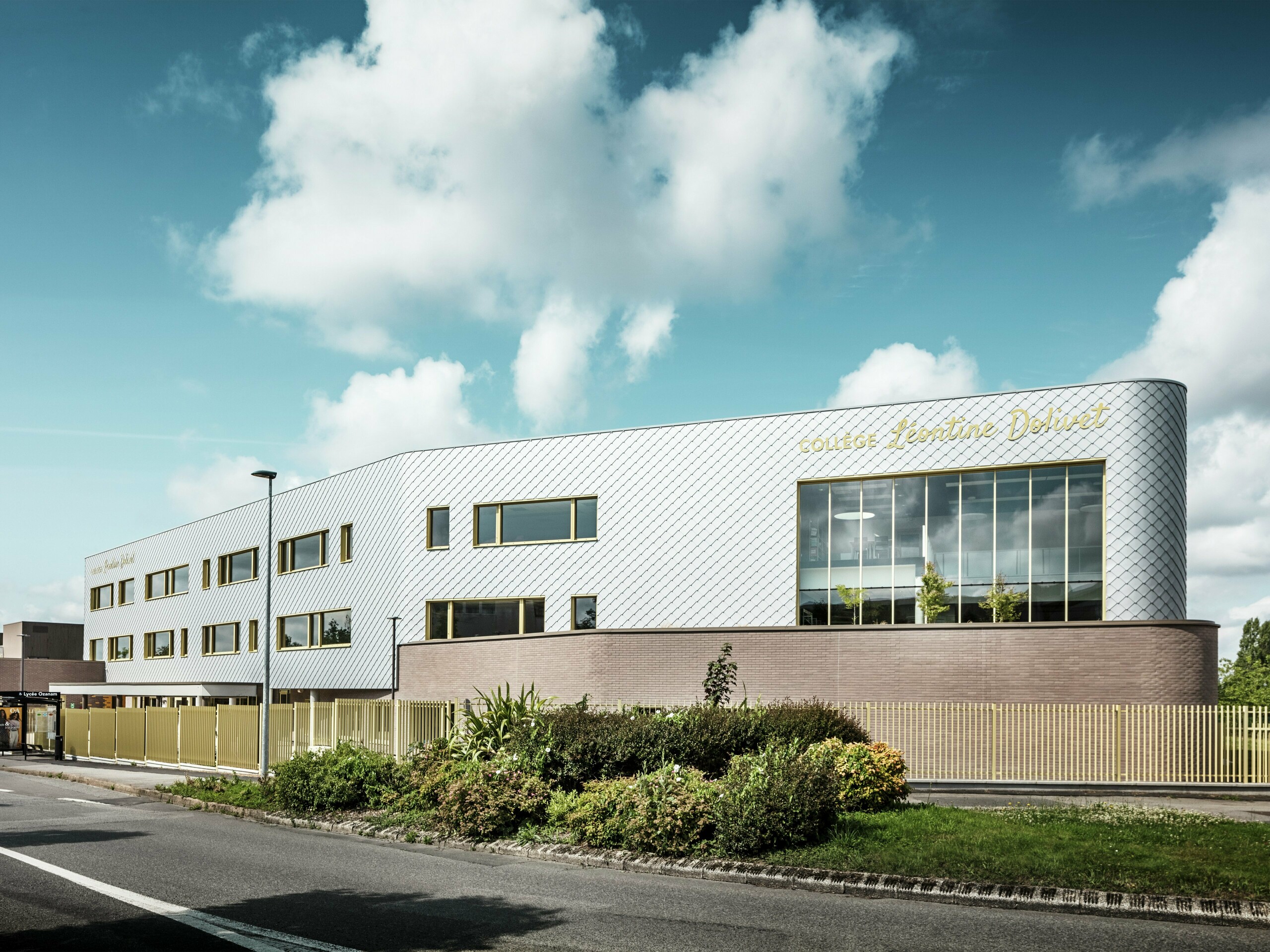 Blick auf das Collège Léontine Dolivet mit seiner markanten, silberglänzenden Aluminiumfassade aus PREFA-Wandelementen (29 × 29 mm) in Silbermetallic. Die Fassade wirkt dank des diagonalen Rautenmusters dynamisch, während die großen Glasflächen und goldenen Fensterrahmen interessante Kontraste bilden. Im Vordergrund sind die gestalteten Grünflächen und der moderne Zaun, der das Gebäude umgibt, zu sehen. Die langlebige PREFA-Fassadenlösung vereint Eleganz, Witterungsbeständigkeit und Nachhaltigkeit – ein architektonisch ansprechendes Gebäude in Cesson-Sévigné bei Rennes.