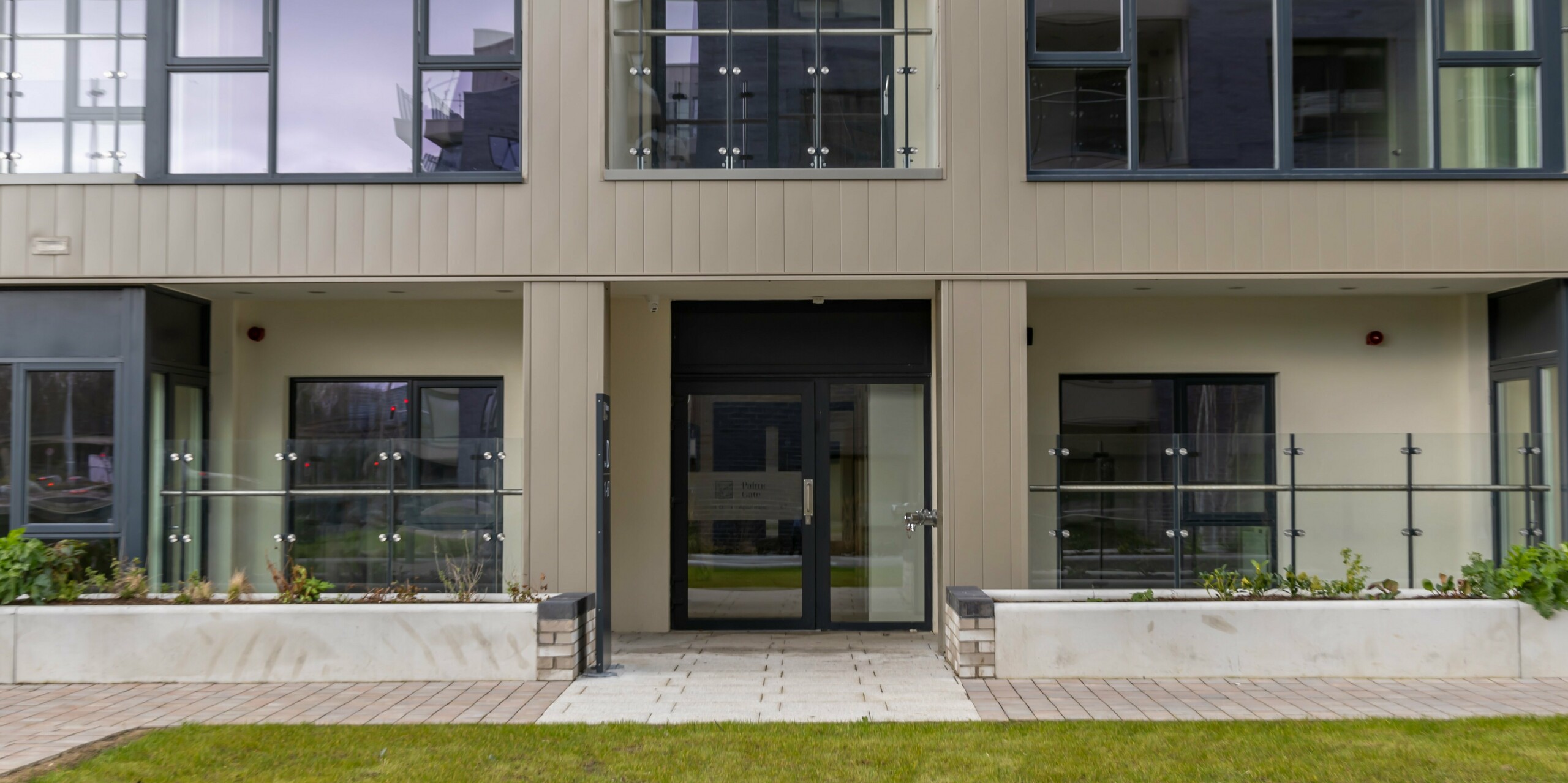 Eingangsbereich des Palmers Gate Wohnkomplexes in Palmerstown, Dublin, Irland. Die Fassade ist mit bronzefarbenen PREFA Sidings verkleidet, die eine moderne und elegante Optik bieten. Der Eingangsbereich ist von großen Glasfenstern umgeben und führt zu den Wohnbereichen. Glasgeländer und Pflanzkästen vor den Fenstern verleihen dem Gebäude zusätzlichen Charme. Die Außenhüllen der Wohnblöcke umfassen insgesamt ca. 1.700 m² PREFA Sidings und ca. 1.200 m² PREFALZ Fassadensystem, die für ihre Robustheit und Langlebigkeit bekannt sind.