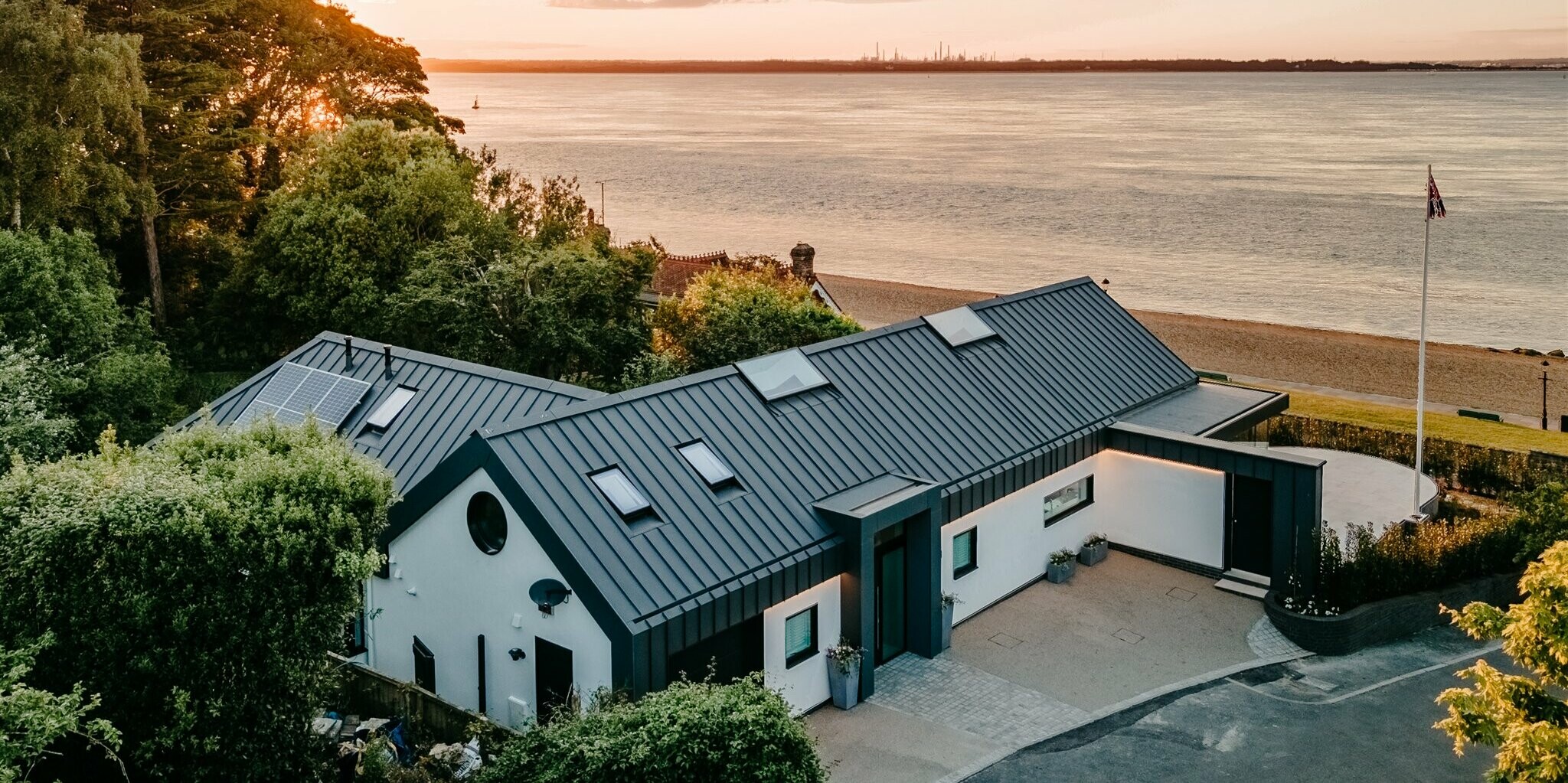 Ein luxuriöser Bungalow auf Isle of Wight, in der Nähe von Bournemouth, wurde mit PREFALZ in der Farbe P.10 Dunkelgrau eingedeckt und fügt sich harmonisch in natürliche Umgebung ein. Das moderne Gebäude mit der eleganten Aluminiumhülle, die mit einer weißen Putzfassade kombiniert wurde, vermittelt ein klares und unaufgeregtes Design. Das architektonische Highlight an der Südküste Englands liegt direkt am Meer. Im Hintergrund wird eine malerische Kulisse in der Golden Hour, der Stunde des Sonnenuntergangs, präsentiert.