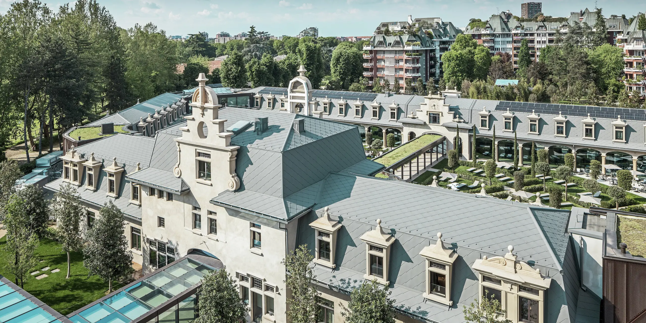 Widok ogólny imponującego kompleksu „De Montel – Terme Milano” z zabytkową architekturą w stylu secesyjnym i nowoczesnym ambientem wellness. Duża powierzchnia pokrycia dachowego z dachówkami PREFA romb 44 × 44 w kolorze P.10 jasnoszarym nadaje kompleksowi elegancką jednolitość. Zintegrowane moduły fotowoltaiczne, zielone dachy i szklane połączenia między budynkami odzwierciedlają dążenie do zrównoważonego rozwoju i nowoczesnego designu w kontekście miejskim.