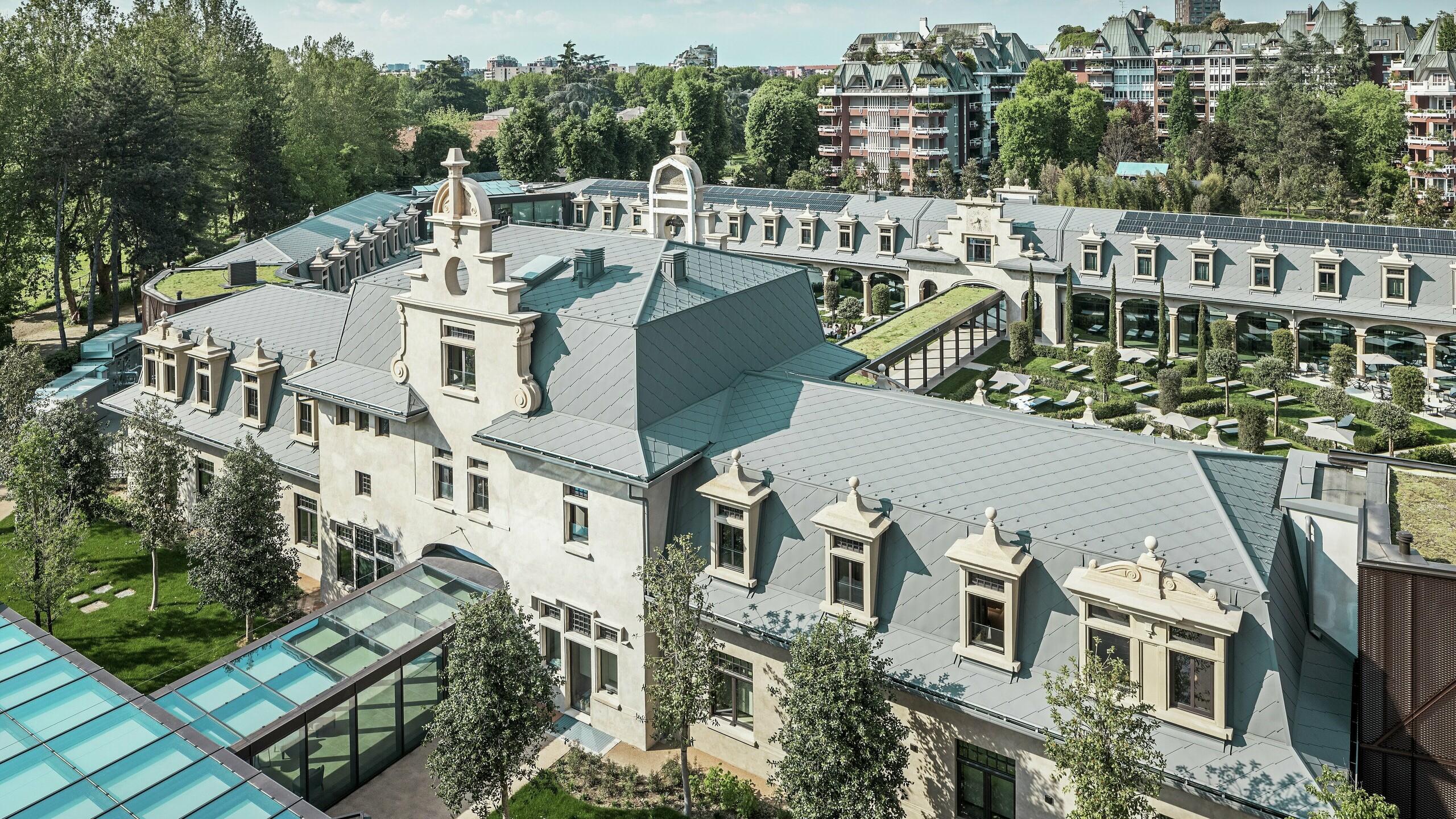 Widok ogólny imponującego kompleksu „De Montel – Terme Milano” z zabytkową architekturą w stylu secesyjnym i nowoczesnym ambientem wellness. Duża powierzchnia pokrycia dachowego z dachówkami PREFA romb 44 × 44 w kolorze P.10 jasnoszarym nadaje kompleksowi elegancką jednolitość. Zintegrowane moduły fotowoltaiczne, zielone dachy i szklane połączenia między budynkami odzwierciedlają dążenie do zrównoważonego rozwoju i nowoczesnego designu w kontekście miejskim.