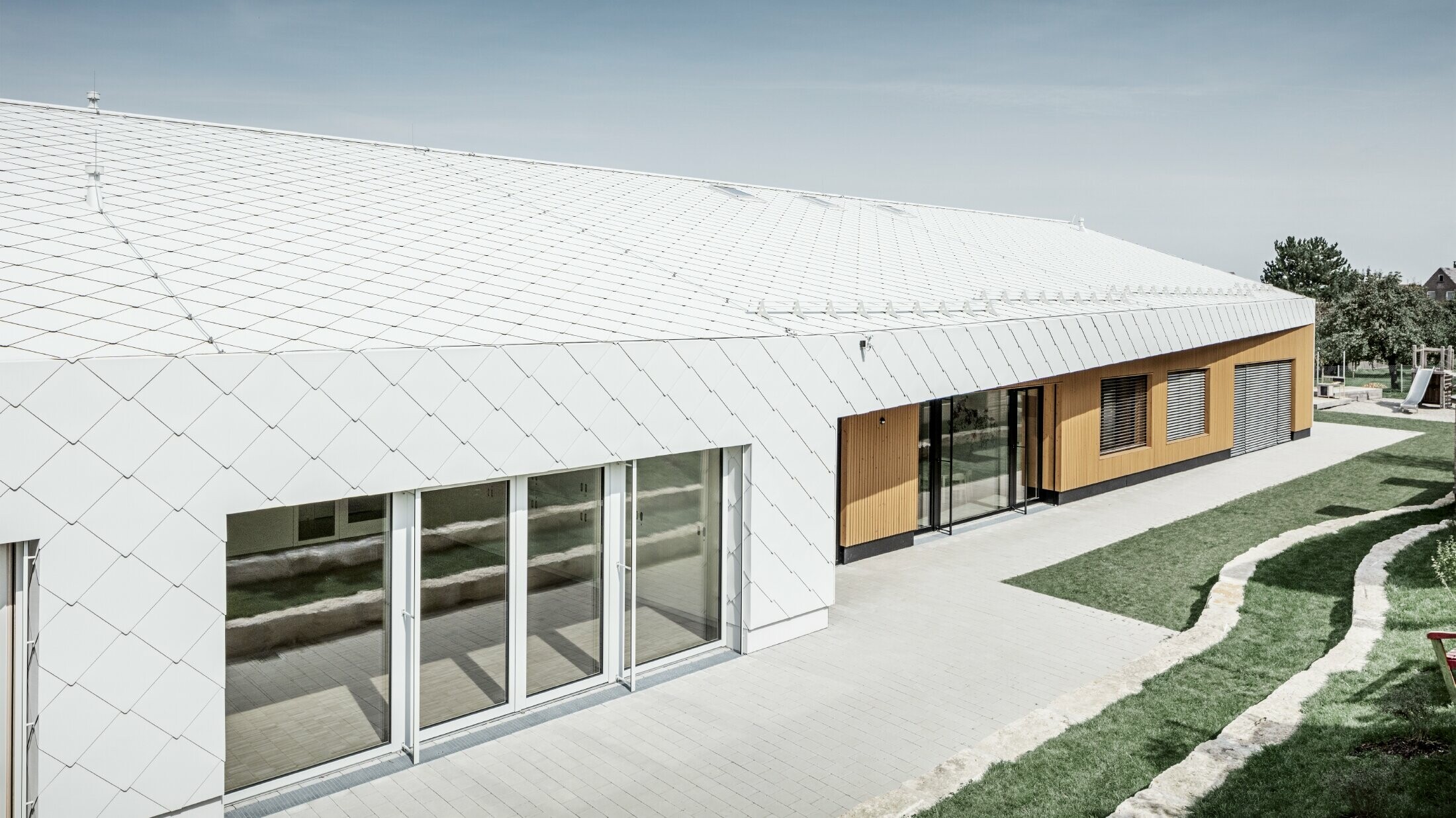 Przedszkole z dachówką romb i rombem elewacyjnym 44 w kolorze białym PREFA. Pokrycie dachu rozciąga się na elewację; duże połacie okien