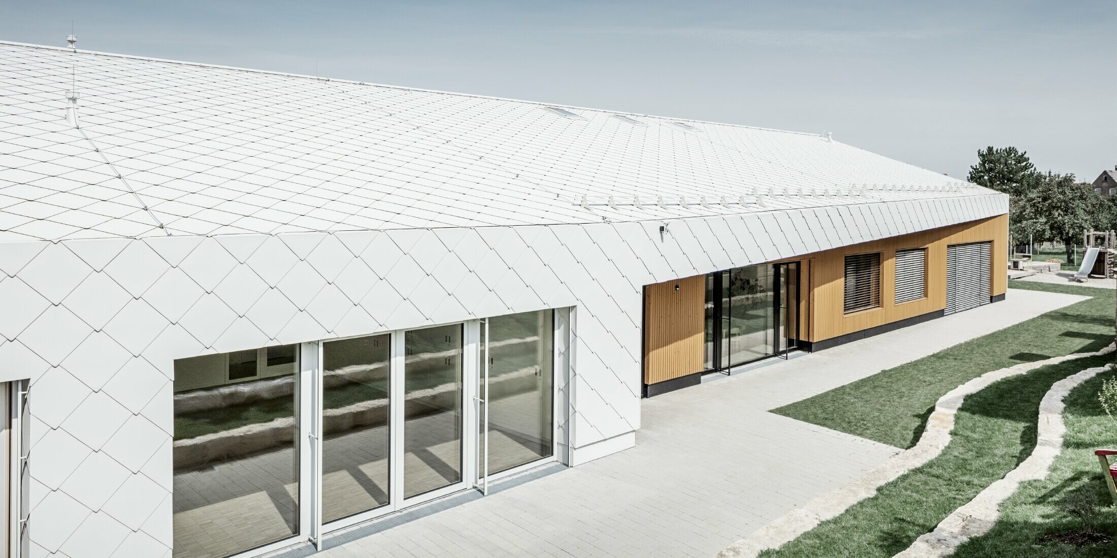 Przedszkole z dachówką romb i rombem elewacyjnym 44 w kolorze białym PREFA. Pokrycie dachu rozciąga się na elewację; duże połacie okien