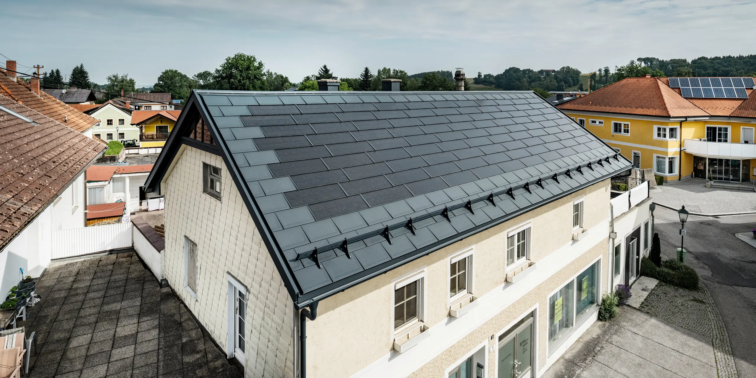 Widok z góry na dom jednorodzinny w Mettmach, Austria. Starszy dom został wyremontowany z użyciem antracytowej dachówki solarnej PREFA. Systemy ochrony przeciwśniegowej i wyraziste linie dachówki solarnej podkreślają precyzyjną obróbkę produktów aluminiowych PREFA. W tle  widoczna jest kontrastująca pomarańczowa dachówka sąsiedniego budynku. Na dachu sąsiedniego domu jednorodzinnego zainstalowane są moduły fotowoltaiczne w klasycznym rozwiązaniu nadachowym, co ilustruje liczne możliwości efektywnego energetycznie projektu domu.