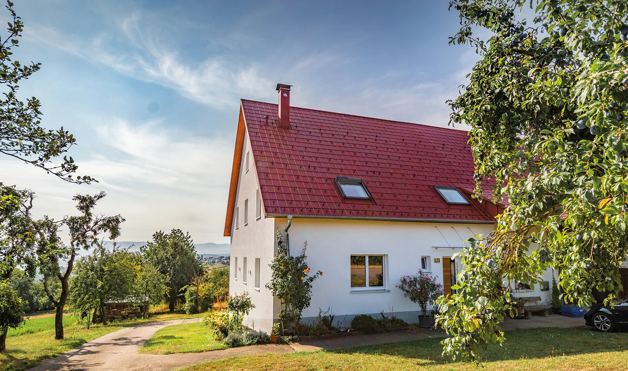 Romantyczny domek na wsi w otoczeniu wielu drzew i krzewów. Pokryty dachem PREFA w kolorze czerwonym tlenkowym.