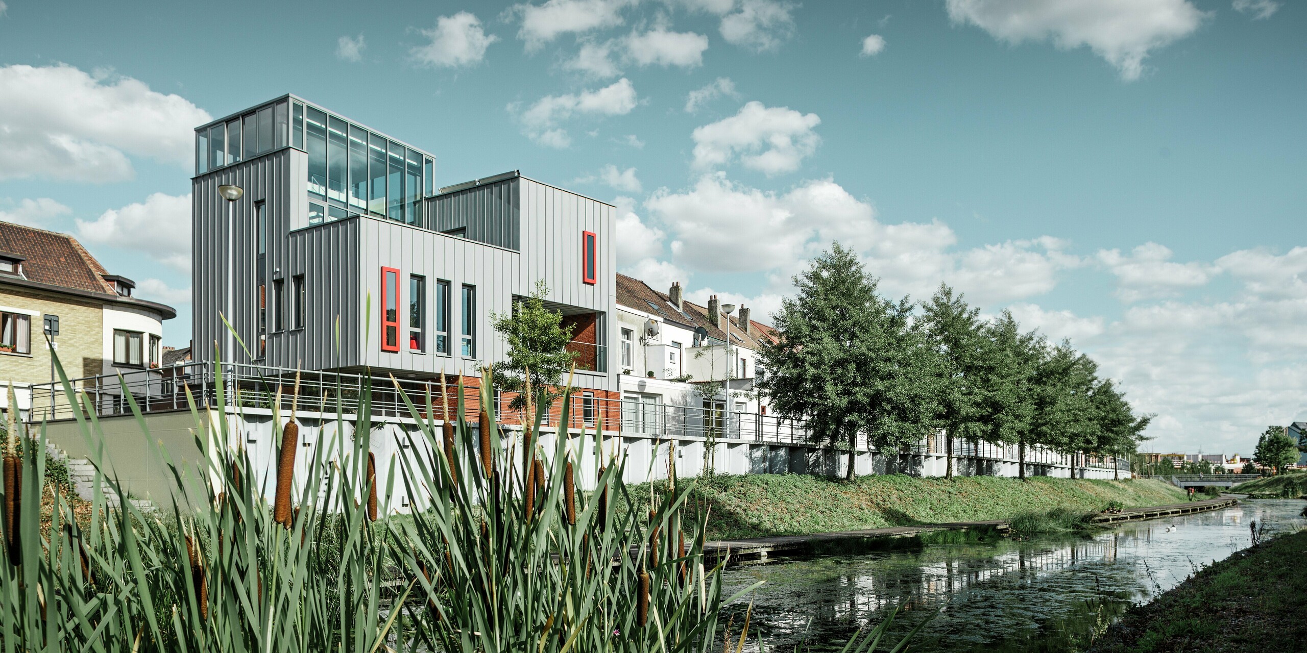 Architektonisches Highlight am Wasser: Das moderne Wohn- und Bürogebäude besticht durch eine PREFALZ Aluminiumfassade in Patinagrau mit vertikaler Stehfalzstruktur. Farblich abgesetzte Fenster in Rot setzen gezielte Akzente. Die Kombination aus Glas, Aluminium und rotem Klinker schafft ein harmonisches Gesamtbild – witterungsbeständig, pflegeleicht und langlebig.