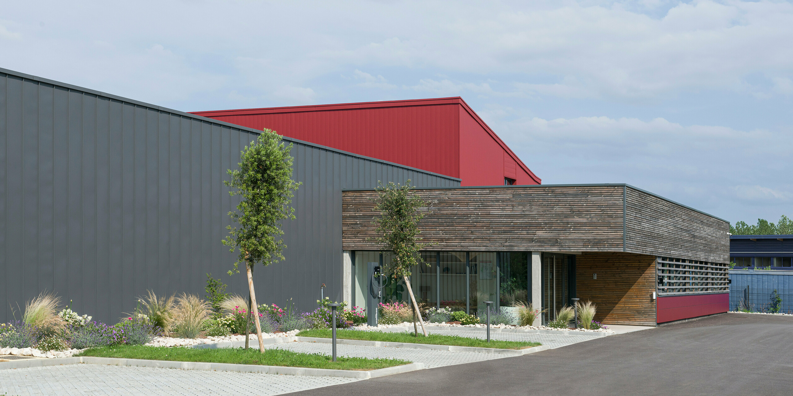 Das Bild zeigt ein modernes Gebäude mit einer auffälligen Kombination aus industriellen und natürlichen Designelementen. Die Fassade kombiniert dunkelgraue und oxydrote Aluminiumelemente, um Dynamik und Textur zu erzeugen. Die oxydroten PREFA Fliesen setzen einen farblichen Akzent. Der untere Teil des Gebäudes mit seiner holzgetäfelten Wand und den großen Fenstern vermittelt eine warme, einladende Atmosphäre, die an einen Büroraum denken lässt. Ein gepflegter Garten vor dem Gebäude vermittelt ein umweltbewusstes und nachhaltiges Image. Die Kombination dieser Elemente spiegelt den Wunsch wider, traditionelle Baupraktiken mit modernen, nachhaltigen Materialien und Designs zu verbinden.