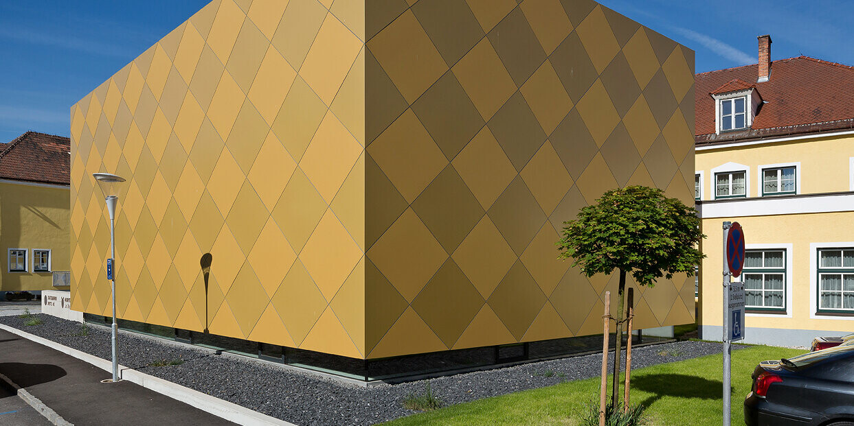 Sicht von der Straße auf den Musikverein in Zwettl. Das quaderförmige Gebäude mit der markanten Außenhülle wurde mit goldfarbenen Aluminium Verbundplatten von PREFA verkleidet. Die karoförmigen Aluminiumelemente unterstreichen das durchdachte Architekturkonzept.