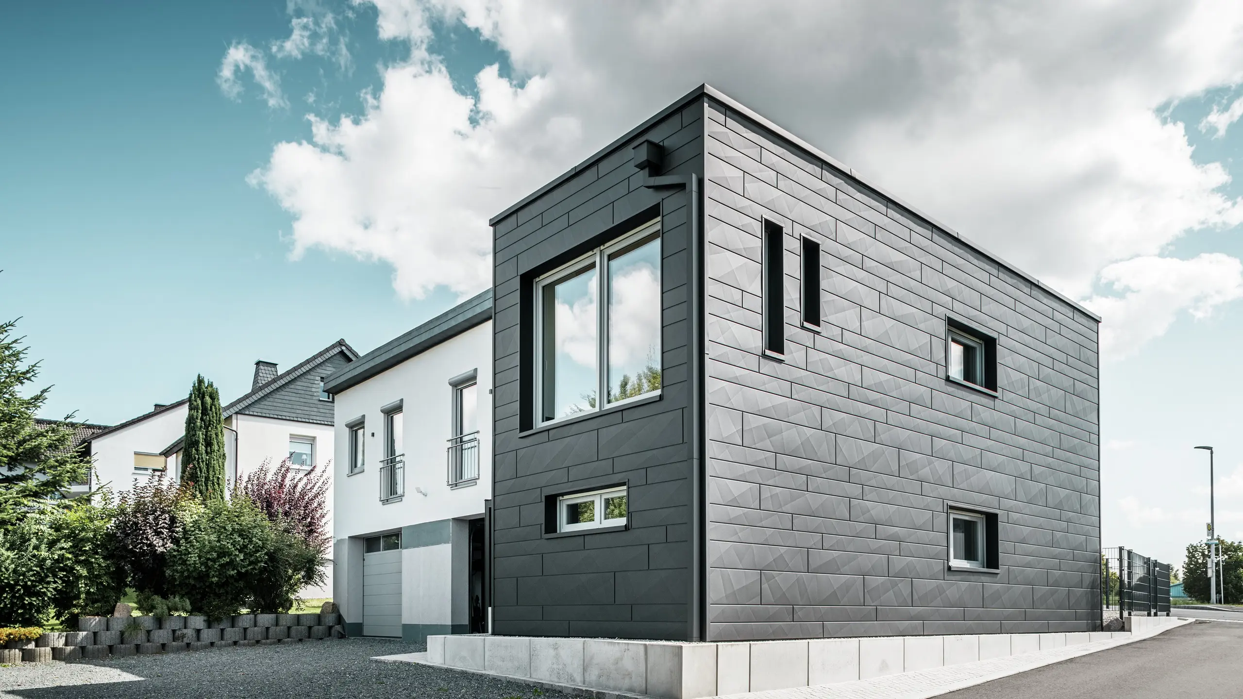 Stylowa i nowoczesna elewacja odnowionego domu jednorodzinnego w Teuschnitz prezentuje panele PREFA Siding.X w kolorze P.10 czarnym o wyraźnej strukturze. Geometryczny układ wysokiej jakości paneli blaszanych nadaje budynkowi charakterystyczną, ponadczasową estetykę. Okna są otoczone czarnymi aluminiowymi płytami kompozytowymi, które harmonijnie dopełniają projekt. Zintegrowane odwodnienie dachu PREFA z rurą kwadratową stanowi funkcjonalne i estetyczne rozwiązanie.