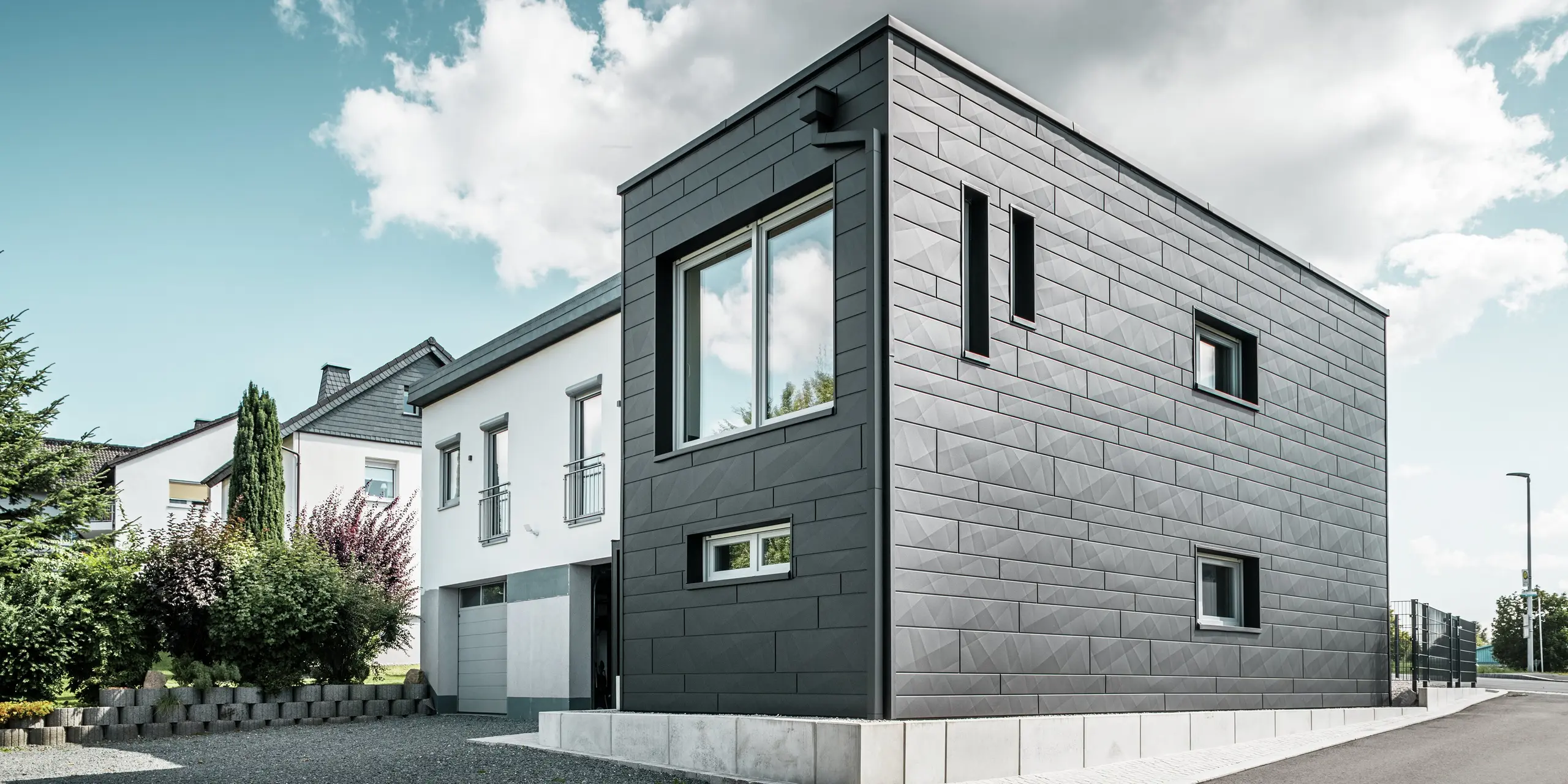Stylowa i nowoczesna elewacja odnowionego domu jednorodzinnego w Teuschnitz prezentuje panele PREFA Siding.X w kolorze P.10 czarnym o wyraźnej strukturze. Geometryczny układ wysokiej jakości paneli blaszanych nadaje budynkowi charakterystyczną, ponadczasową estetykę. Okna są otoczone czarnymi aluminiowymi płytami kompozytowymi, które harmonijnie dopełniają projekt. Zintegrowane odwodnienie dachu PREFA z rurą kwadratową stanowi funkcjonalne i estetyczne rozwiązanie.