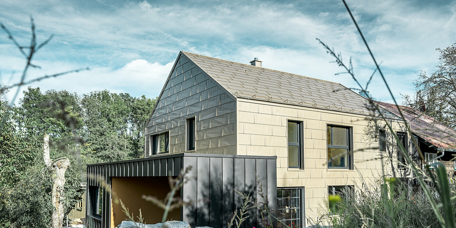 Budynek mieszkalny bez okapu, wykończony na dachu i elewacji aluminiowym panelem PREFA FX.12 w kolorze medalowy brąz Bezpośrednio obok stoi garaż z dachem płaskim z elewacją na zaczep kątowy stały.