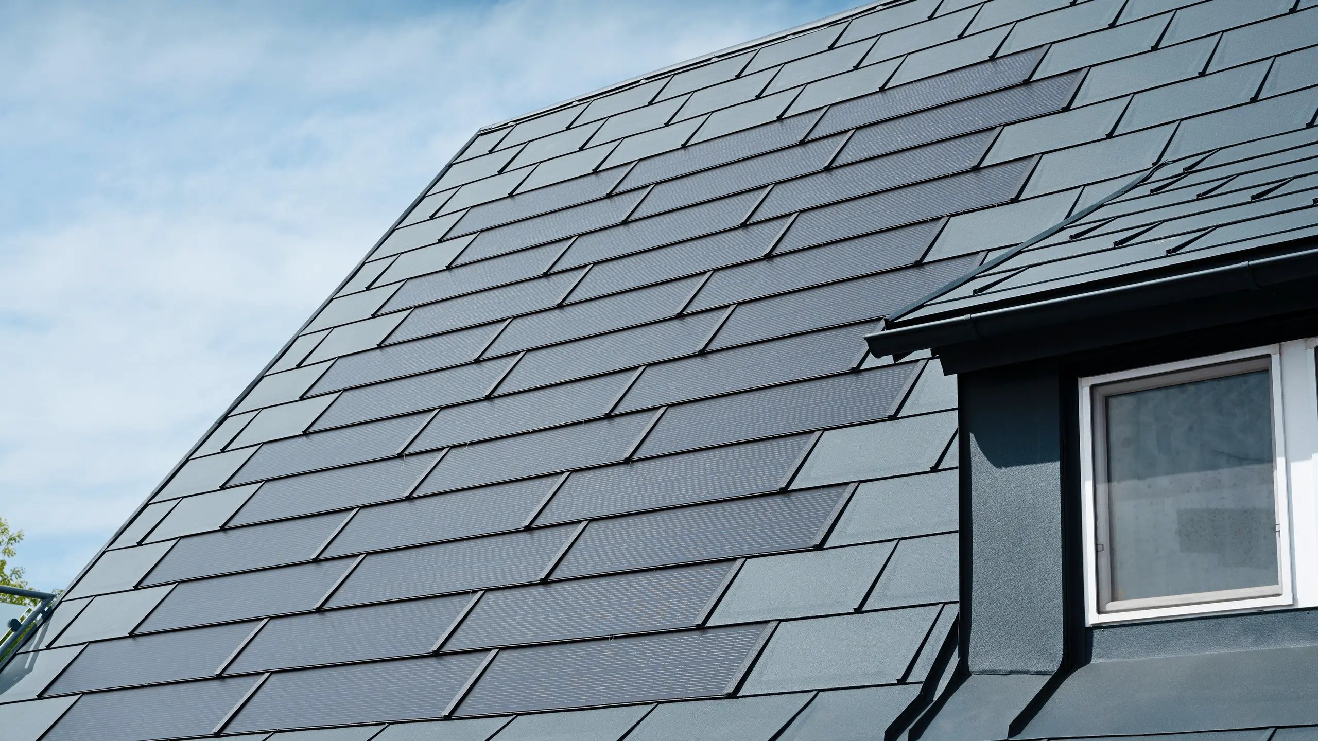 Widok szczegółowy wyremontowanego dachu domu jednorodzinnego w Leonding z wykorzystaniem dachówki solarnej PREFA w kolorze antracytowym. Na zbliżeniu widoczne są wysokiej jakości płyty aluminiowe PREFA, umożliwiające spójną integrację modułów solarnych. To trwałe, odporne na warunki atmosferyczne rozwiązanie zapewnia zrównoważoną, efektywną energetycznie możliwość generowania prądu. Na zdjęciu widoczna jest estetyczna i funkcjonalna jakość zastosowanych materiałów. Przy prawej krawędzi zdjęcia widać białe okno pod lukarną.